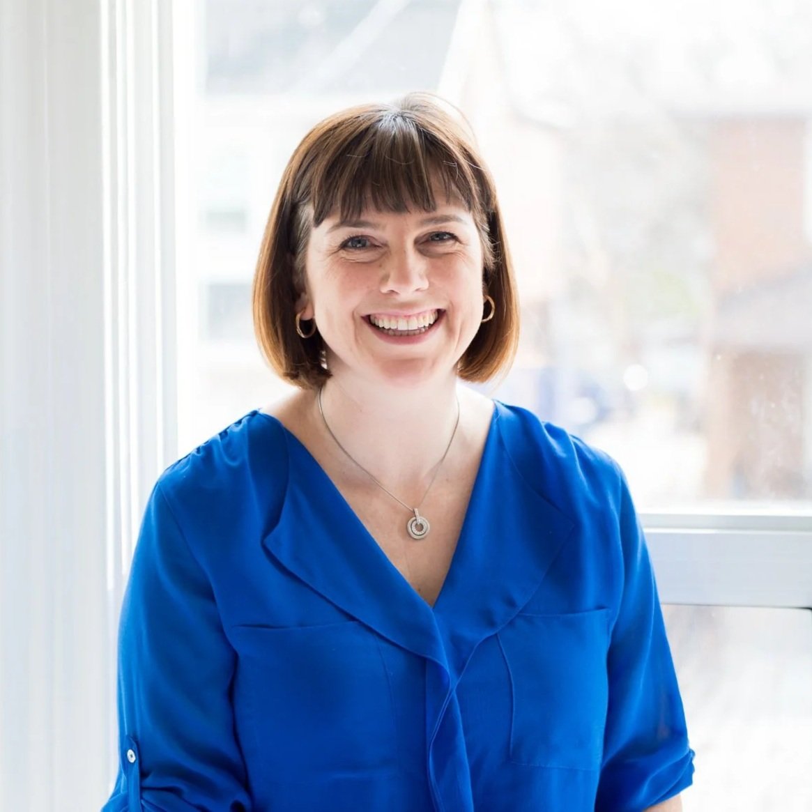 A woman with short brown hair and light skin, smiling and wearing a blue blouse, sitting near a window with bright natural light.