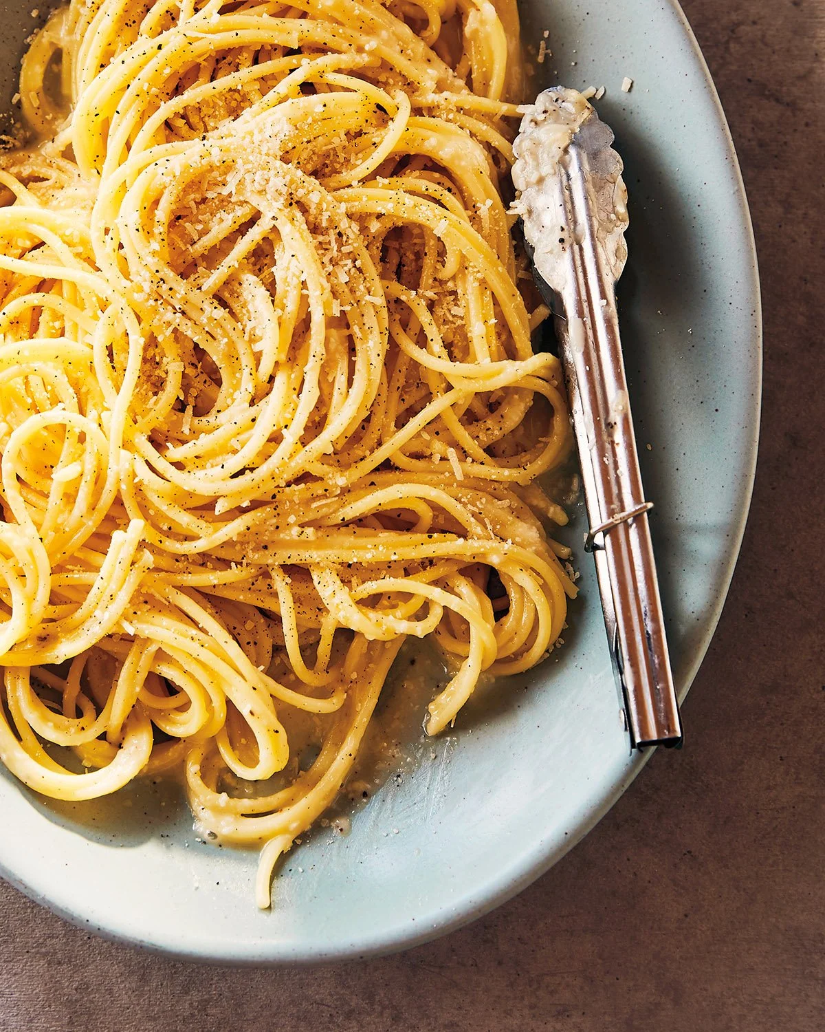 A plate of spaghetti pasta sprinkled with grated cheese and black pepper, with a metal pasta serving spoon on the right side.