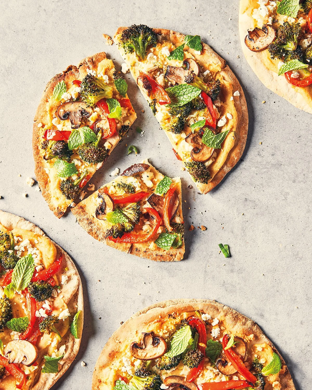 Multiple slices of vegetable pizza with broccoli, mushrooms, red bell peppers, and mint leaves on a grey surface.