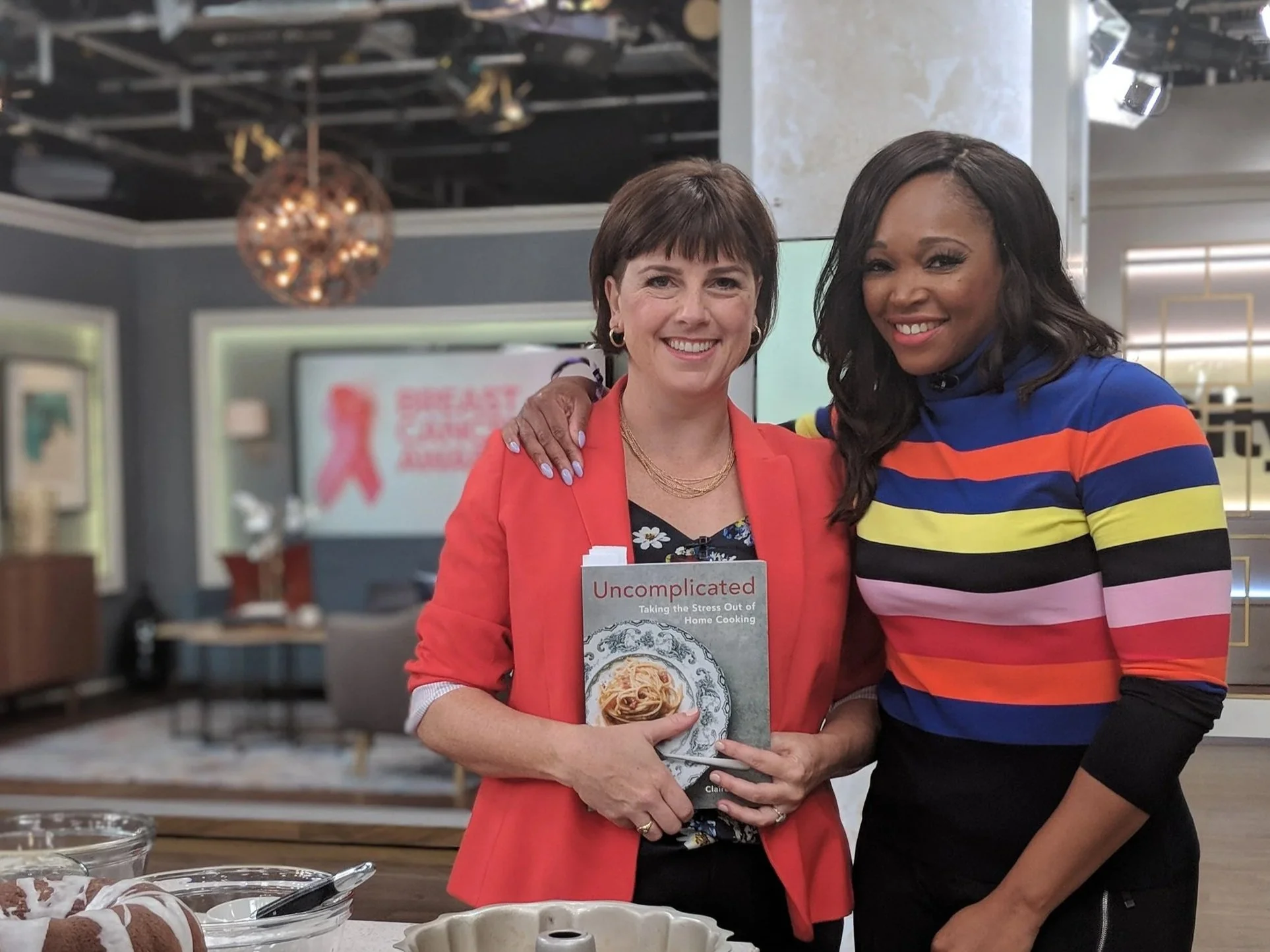 Two women smiling and posing for a photo in a kitchen or cooking studio. One woman holds a cookbook titled "Uncomplicated" with a picture of a pasta dish on the cover. The woman on the left has short brown hair, wearing a red blazer and floral blouse, and the woman on the right has long dark hair, wearing a colorful striped top. There are baking ingredients and kitchen tools on the counter in front of them.
