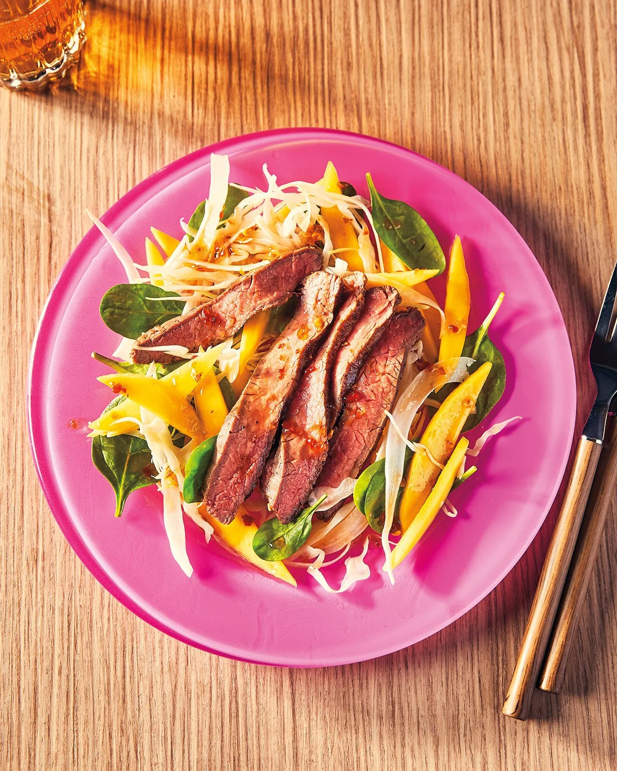 A pink plate with sliced grilled steak on a bed of mixed greens and vegetables, on a wooden table with chopsticks and a glass of amber-colored beverage.
