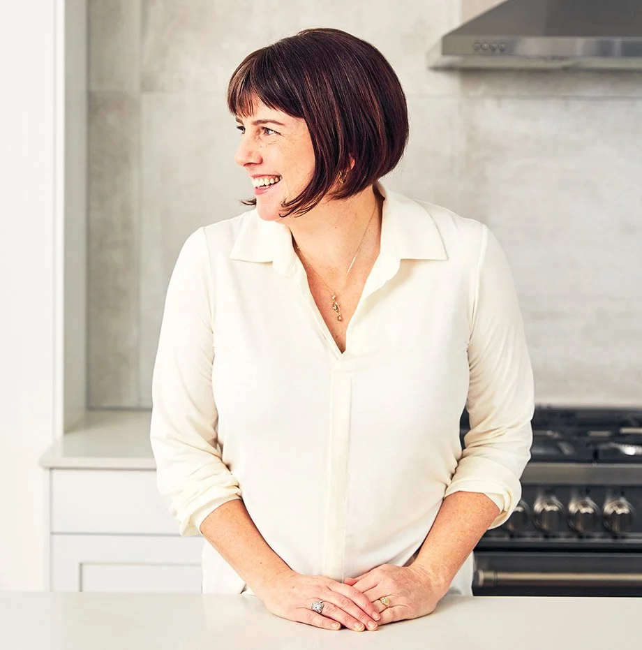 A woman with short brown hair and light skin, smiling and looking to her left, standing in a modern kitchen with a stove behind her, wearing a white blouse and ring on her finger.