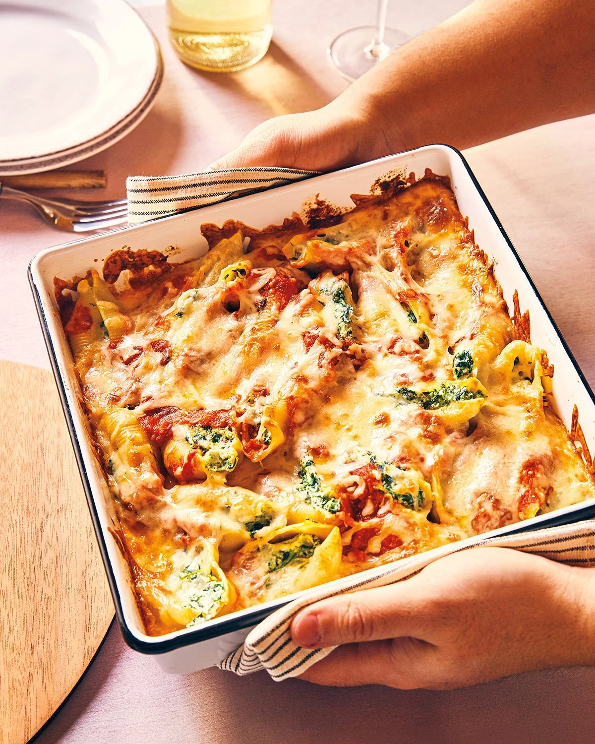 Person holding a freshly baked cheesy pasta casserole in a white baking dish.