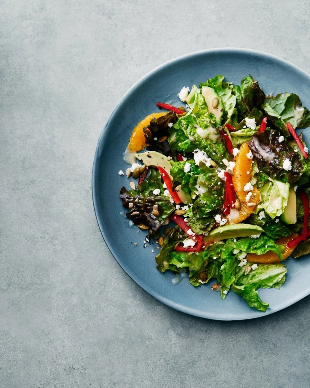 A bowl of mixed green salad with red peppers, orange slices, avocado, seeds, and crumbled cheese on a gray background.