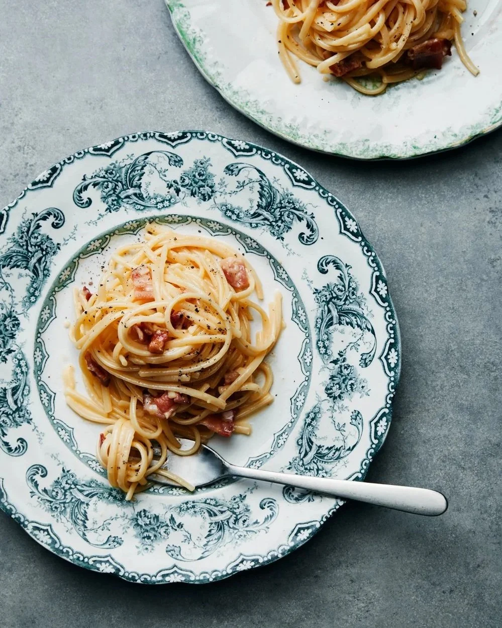 Plate of spaghetti with bacon on a decorative floral plate, with another plate of spaghetti in the background.