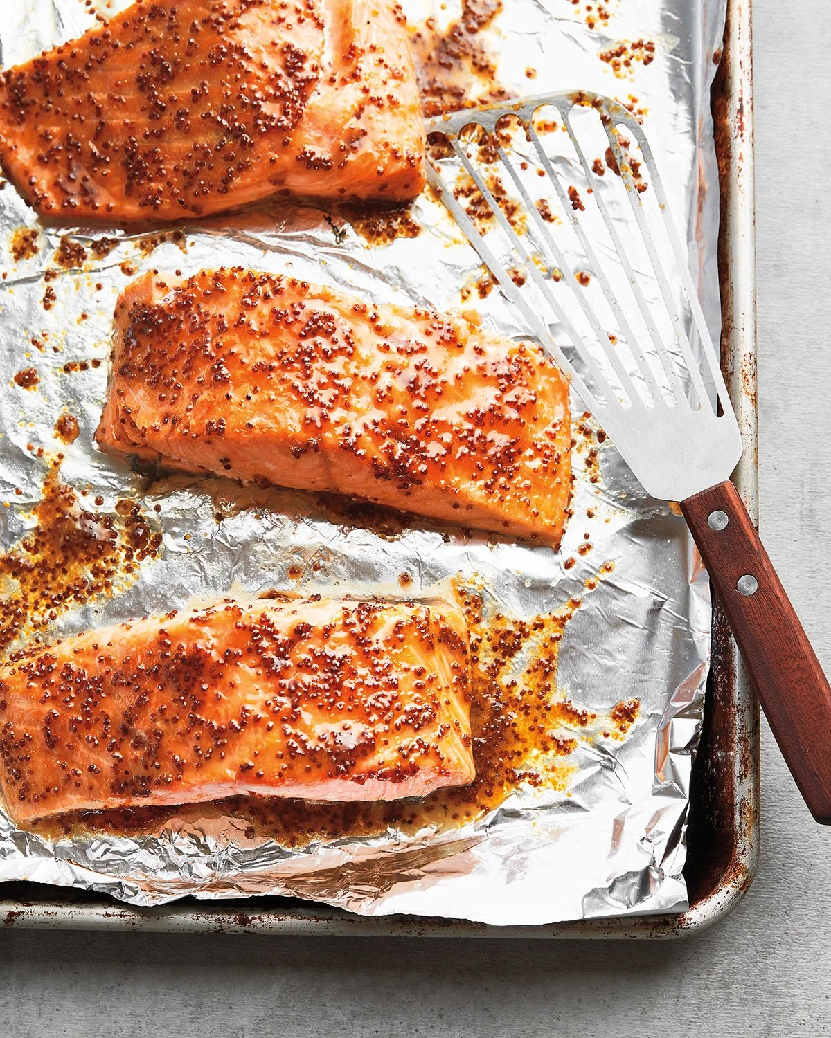 Baked salmon fillets sprinkled with mustard seeds on a foil-lined baking sheet, with a stainless steel spatula.