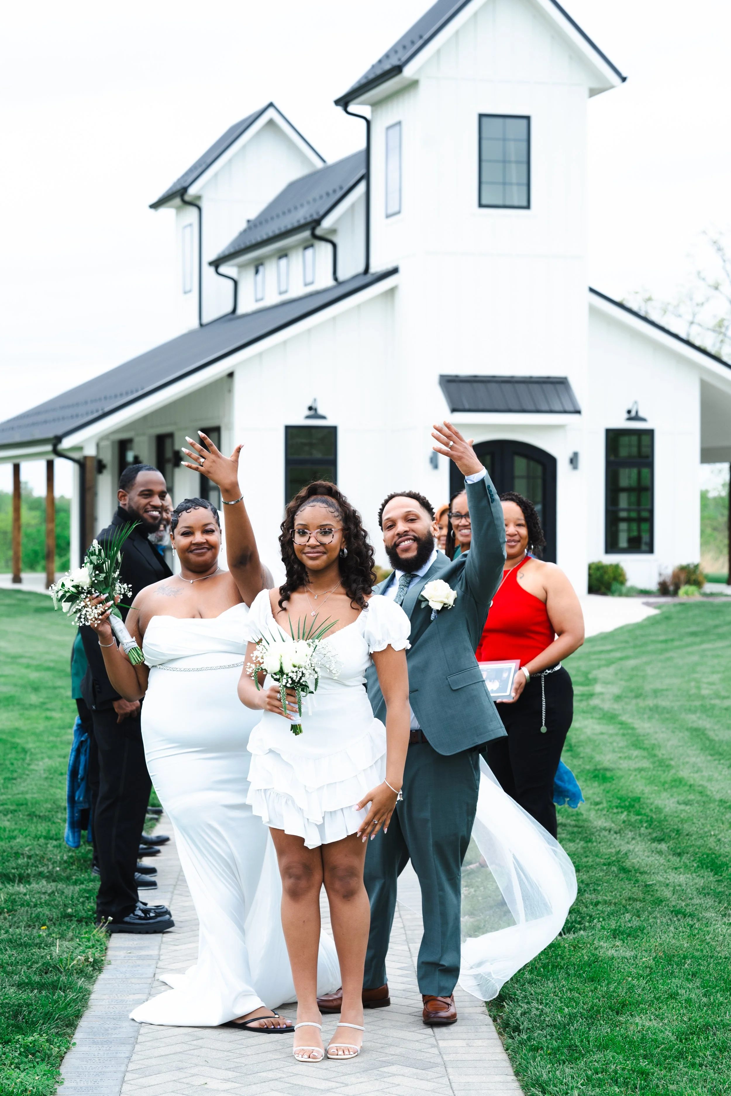 A wedding party stands on a sidewalk in front of a white house with a black roof, celebrating outdoors. The bride is in the center wearing a white dress, holding a bouquet, and smiling. The groom is next to her, wearing a gray suit, raising his hand, and smiling. Bridesmaids and groomsmen, dressed formally, stand around them, some smiling and waving.
