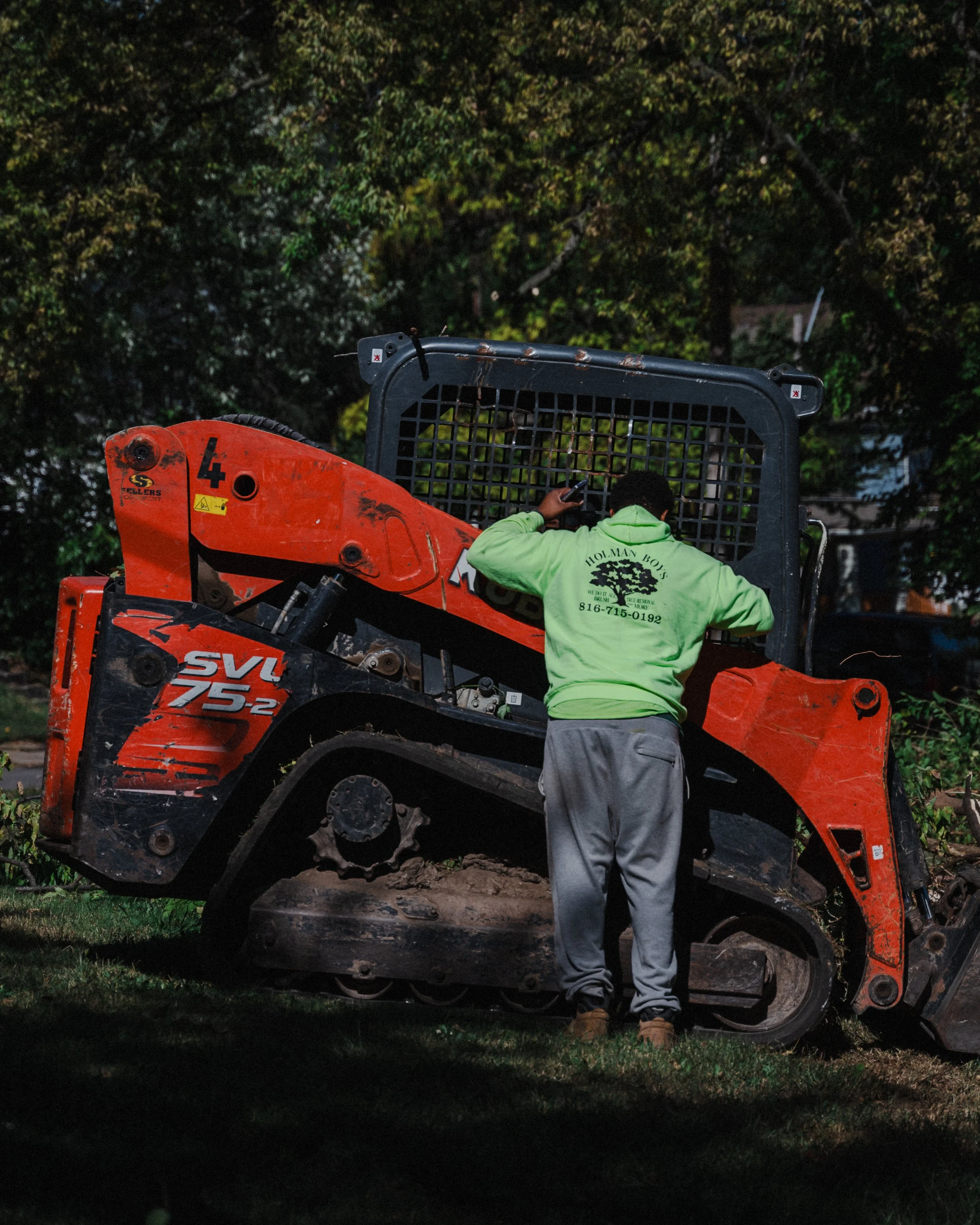 A person in a green hoodie and gray pants operating or inspecting a small orange and black skid-steer loader in a wooded area during evening or night.