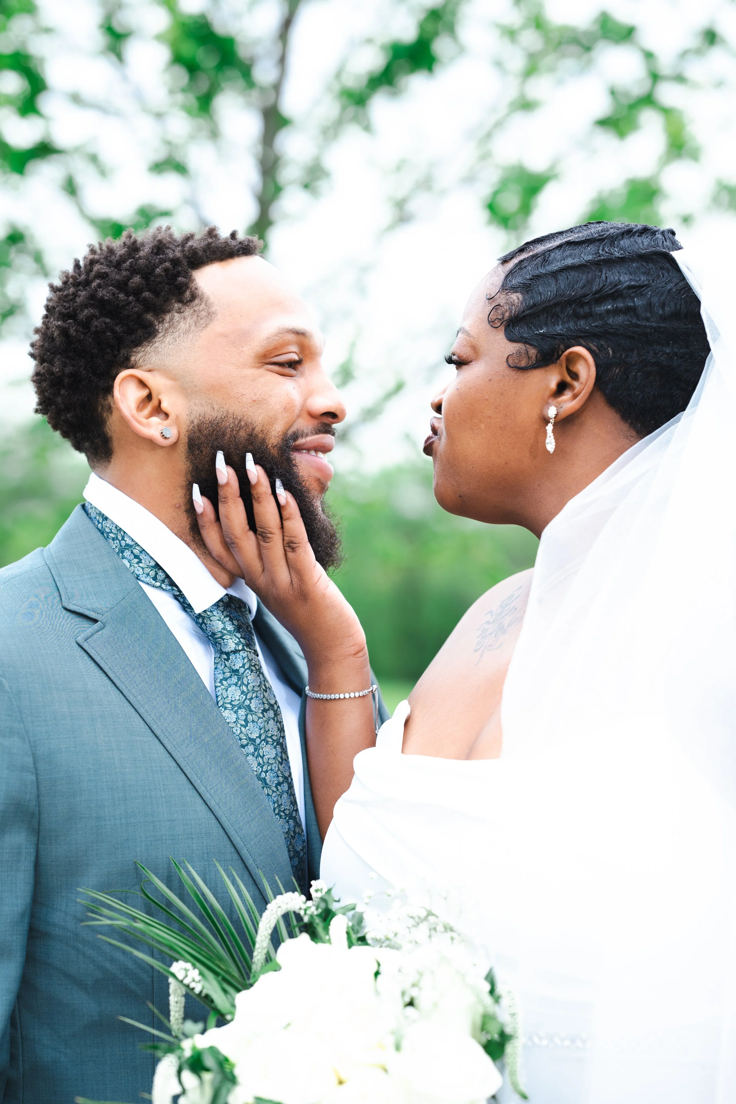 A couple on their wedding day, a man in a teal suit and a woman in a white wedding dress, standing face-to-face outdoors with greenery in the background.