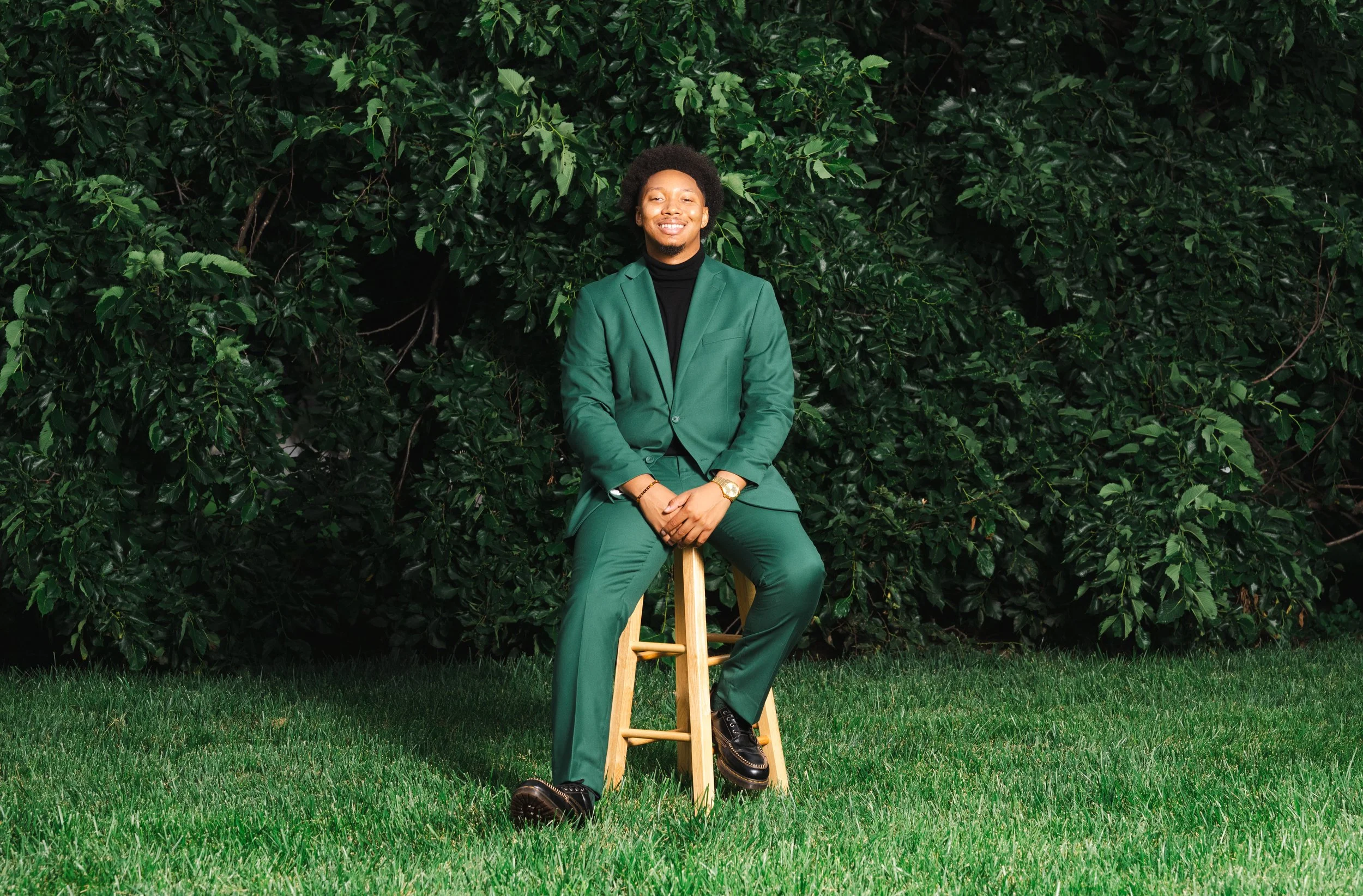 A young man in a green suit sitting on a wooden stool in front of a dense leafy green hedge, smiling at the camera.