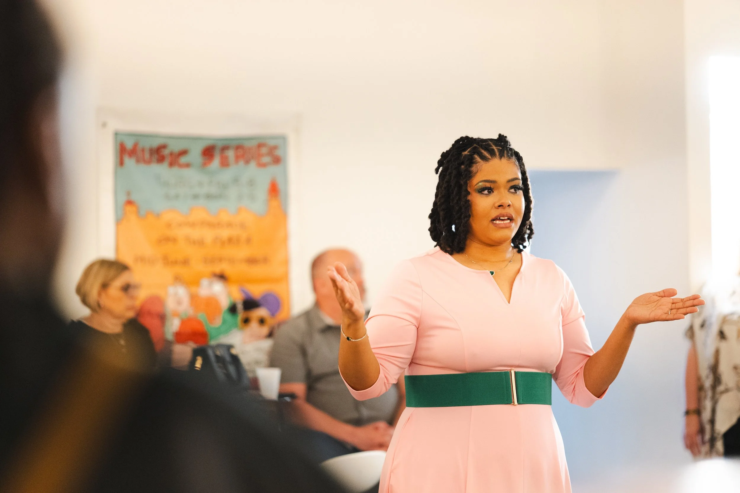 A woman with dark, curly hair styled in braids and wearing a pink and green dress is speaking and gesturing with her hands during a presentation. In the background, there are seated audience members and a poster that reads 'Music Spaces'.