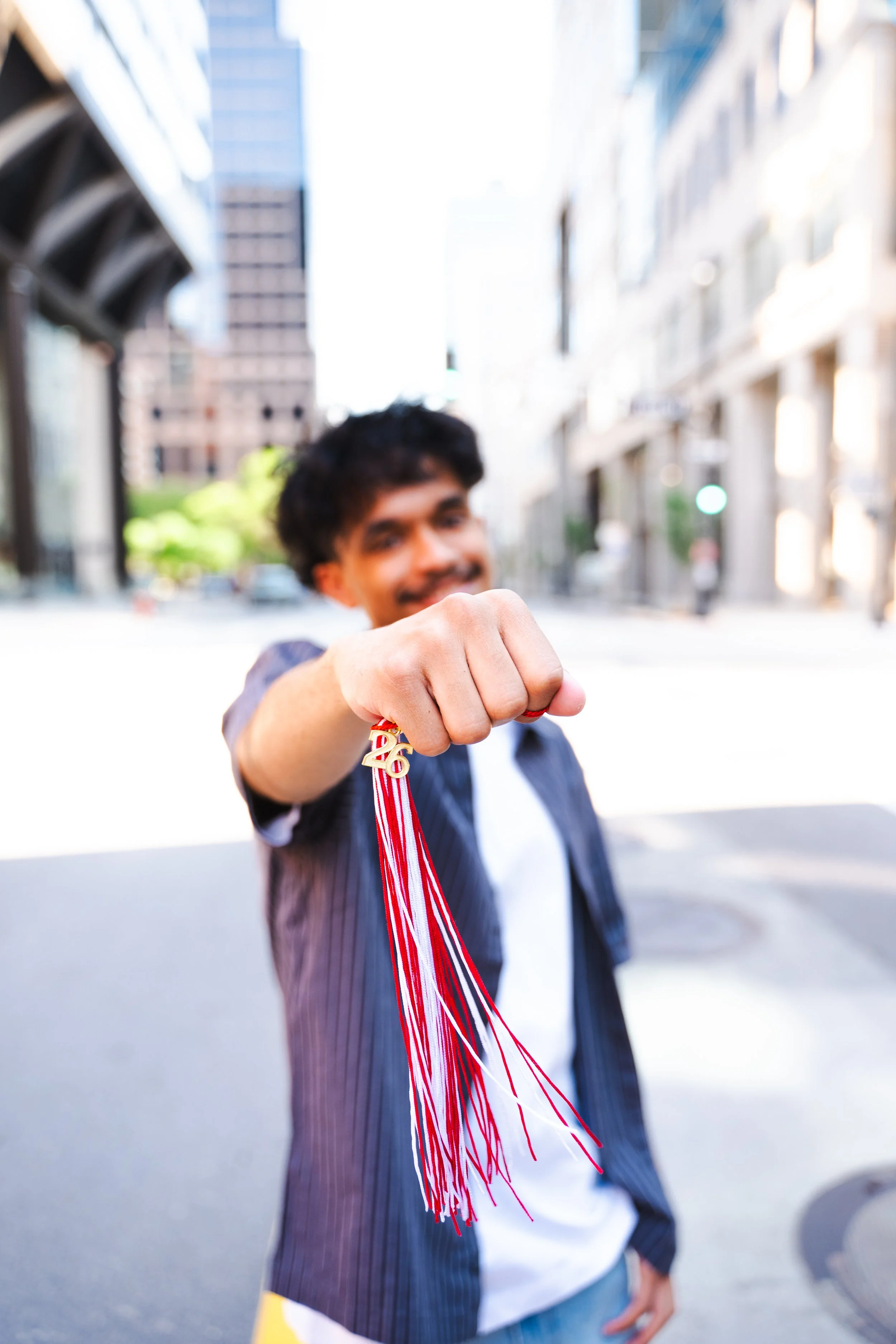 Smiling young man in a suit holding a gold medal with red and white ribbon, standing outdoors in an urban setting with buildings and a street in the background.