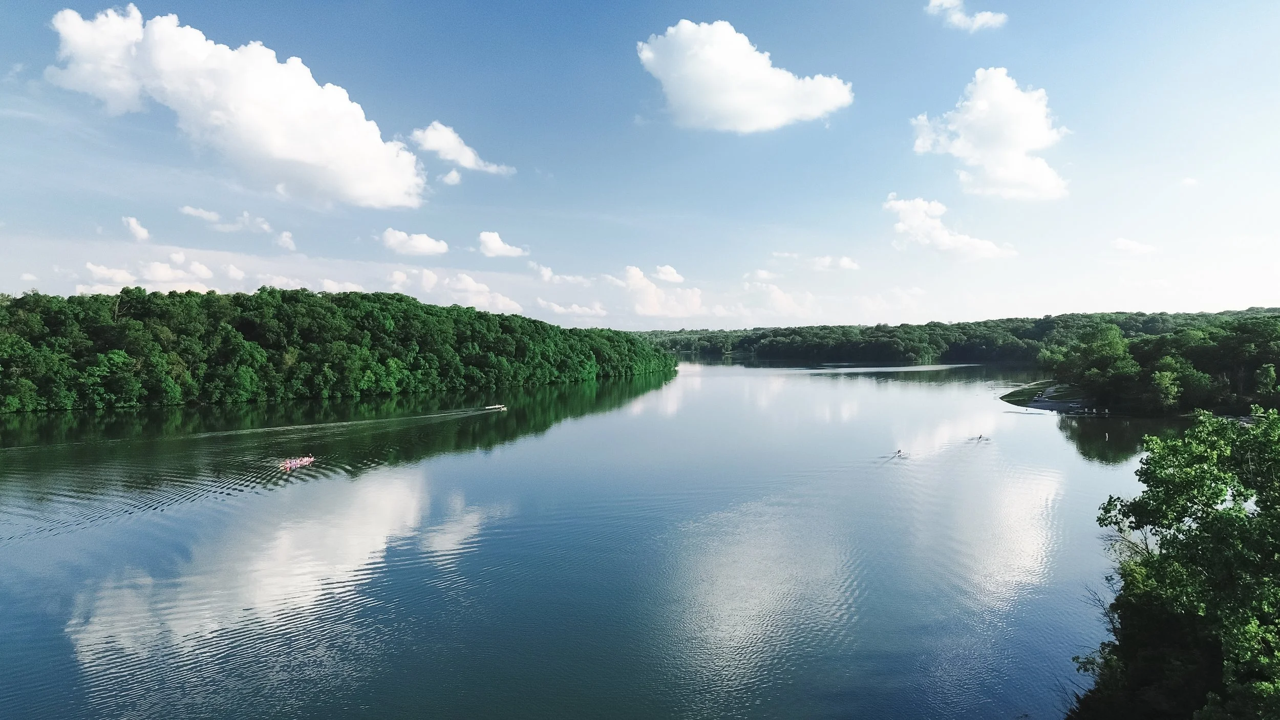 A calm river flowing through a lush green forest, with a few boats and kayaks on the water under a bright blue sky with fluffy white clouds.