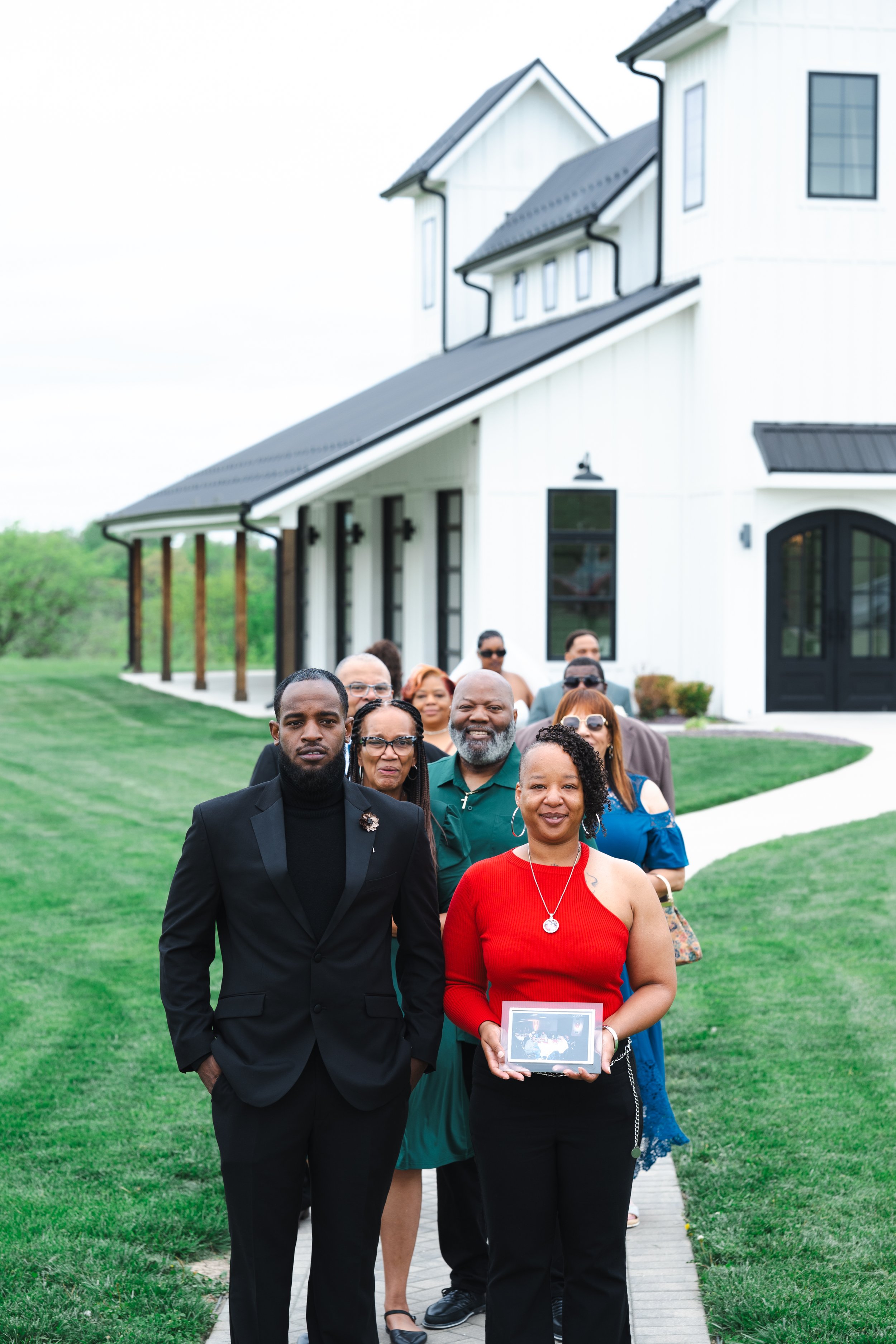 A group of diverse people standing in line on a sidewalk in front of a white modern house with black trim, during daytime, on a green lawn.