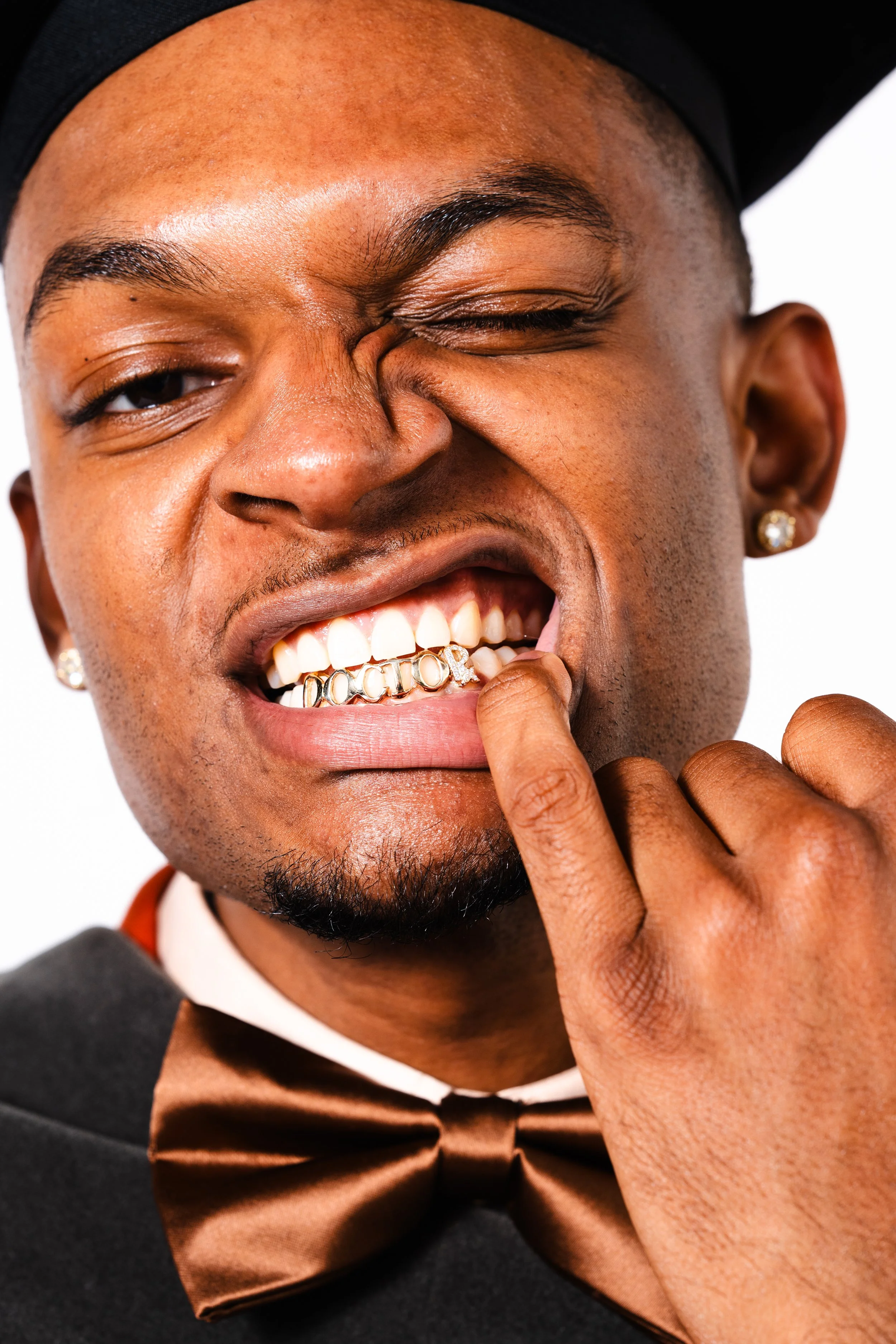 A young man with a dark skin tone smiling and winking. He is wearing a tuxedo with a bow tie, earrings, and has gold teeth. He is pulling down his lower lip with one finger.