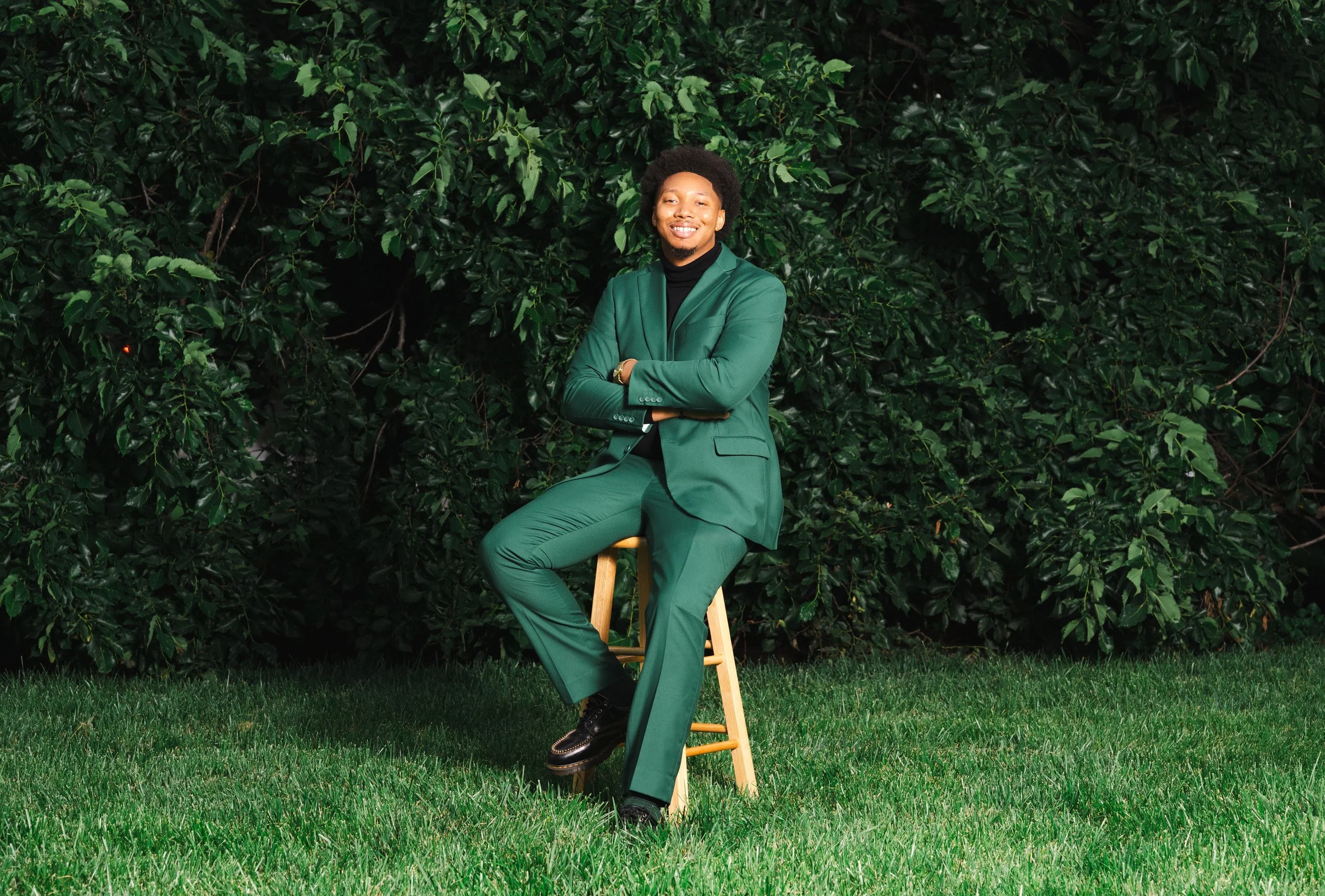 A man with an afro hairstyle wearing a green suit and black turtleneck, smiling with arms crossed, sitting on a wooden stool in front of a background of dense green foliage.