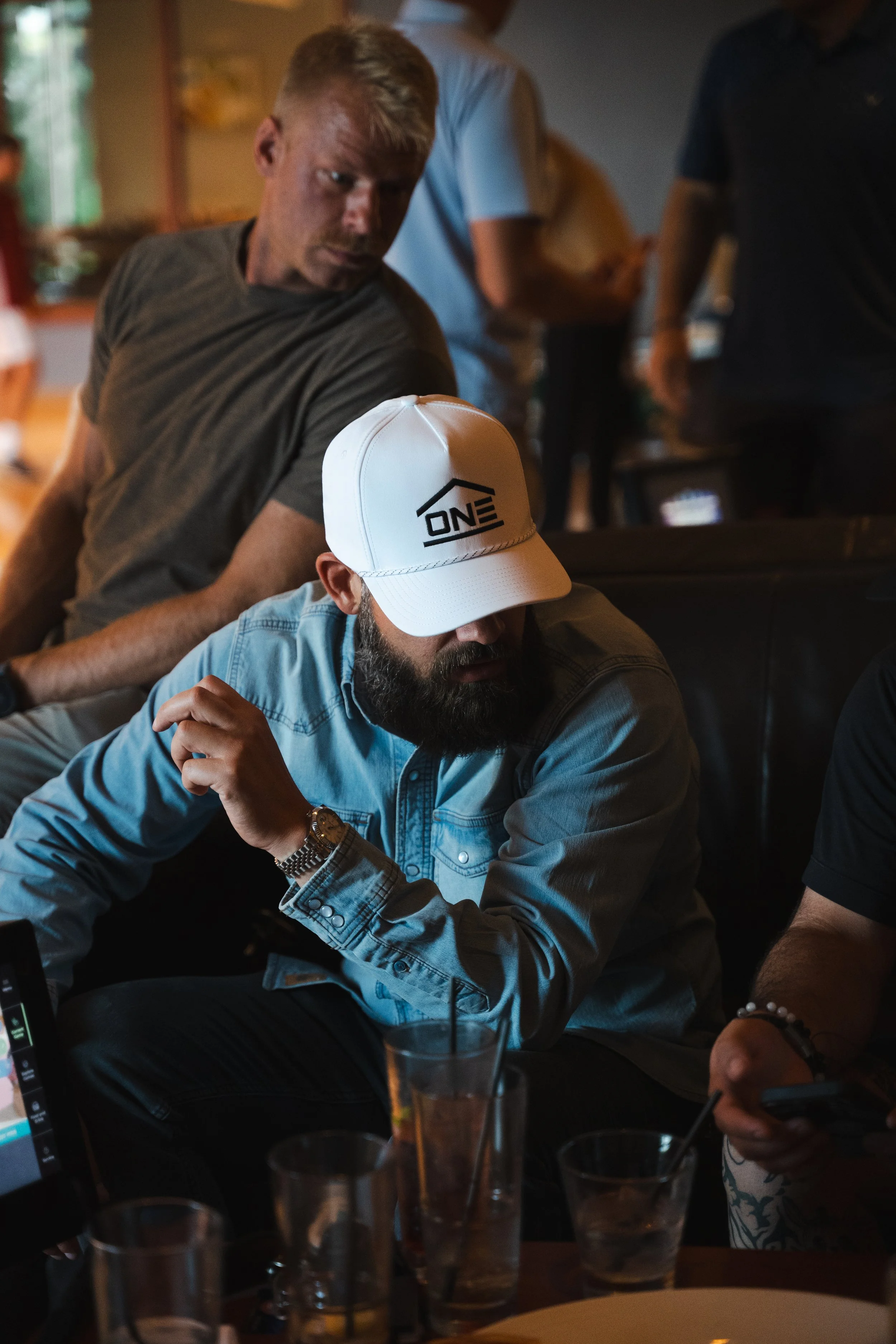 A man with a beard wearing a light blue denim shirt and a white cap with the logo 'ONE,' sitting in a dimly lit restaurant. Several drinks in glasses are on the table in front of him. Other people are in the background, standing or sitting, engaged in conversation.