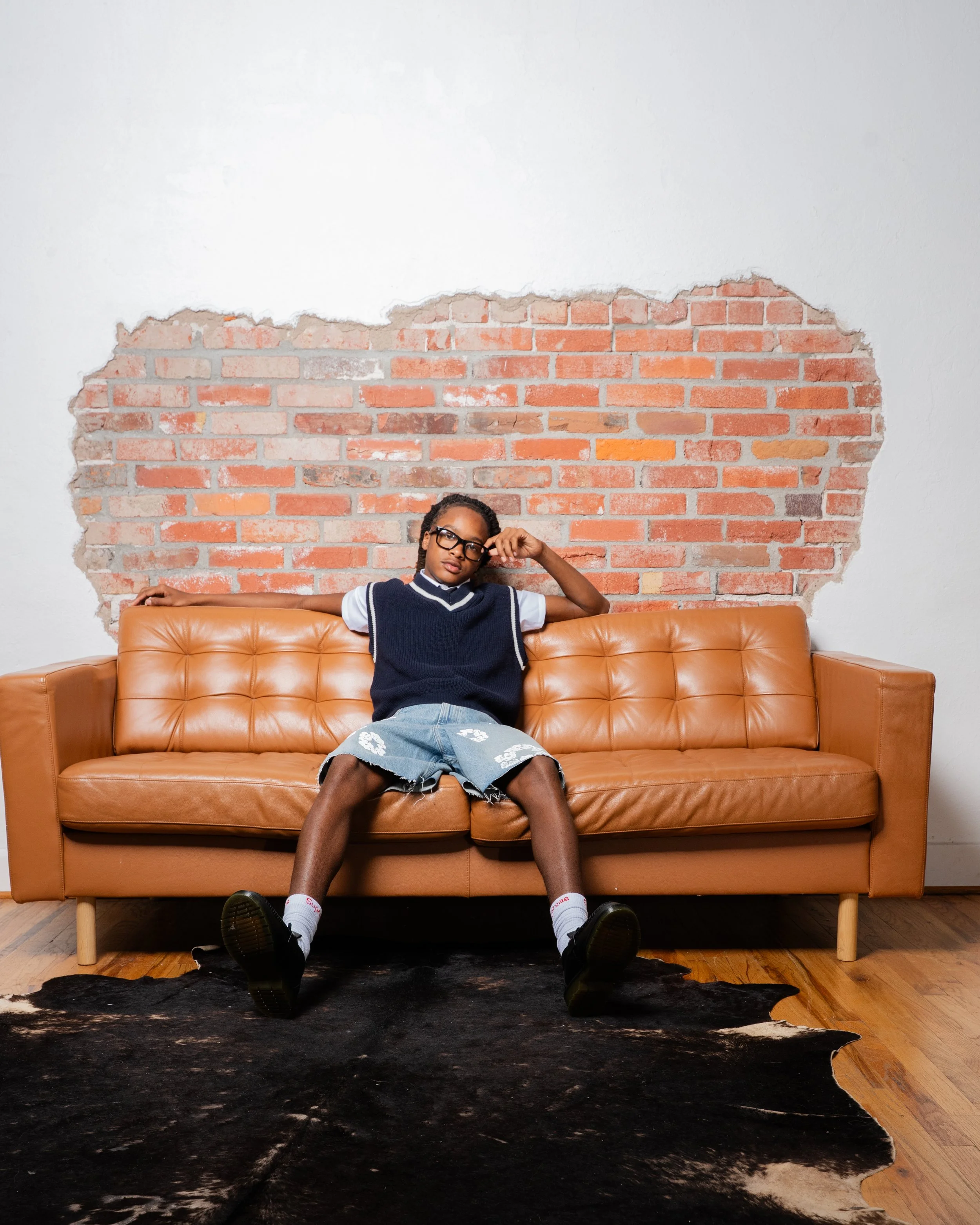 A person with glasses and braided hair poses on a leather couch in front of a partially exposed brick wall.