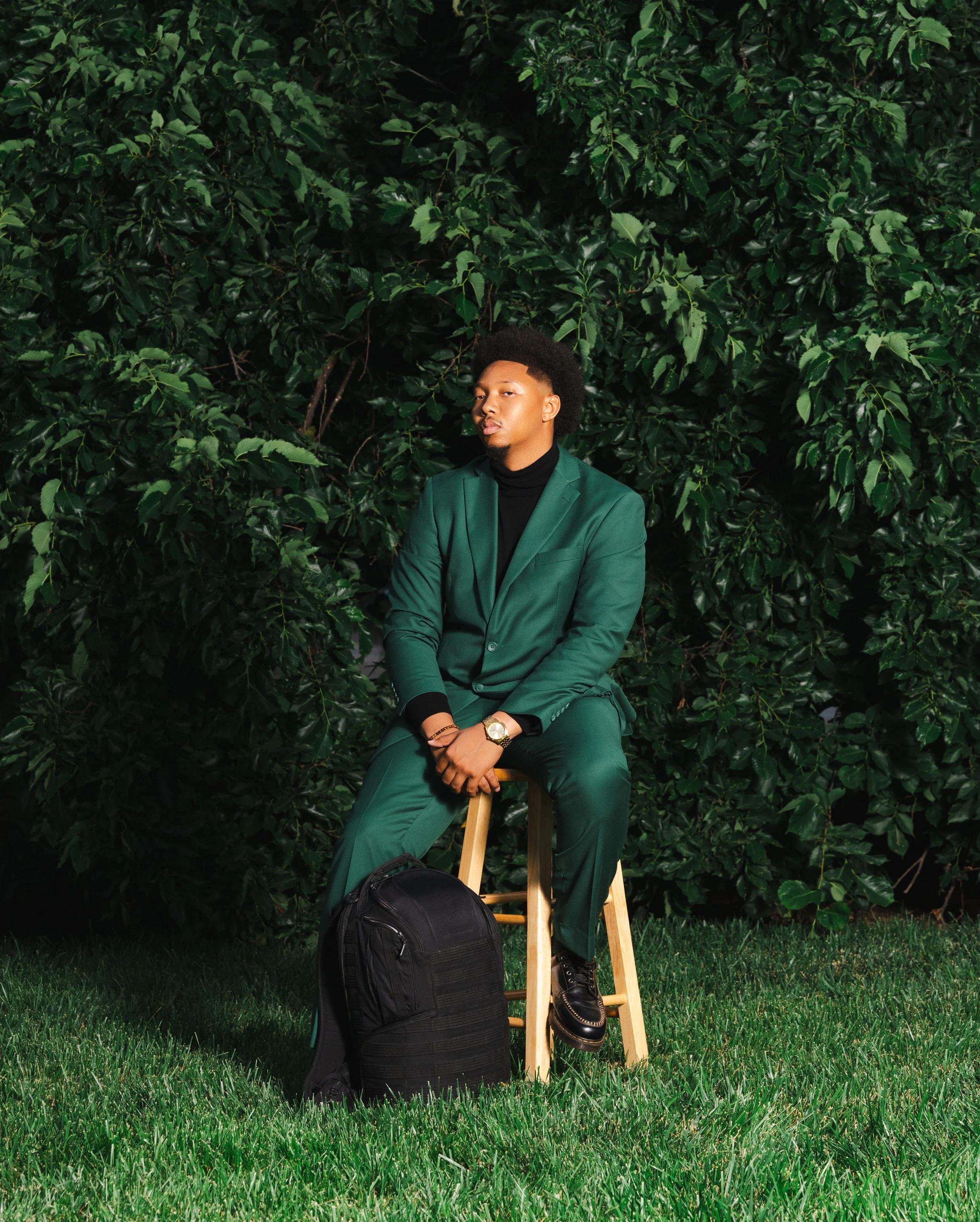 A young man with black hair and a goatee wearing a green blazer and matching pants, black turtleneck shirt, sitting on a stool in front of a leafy green hedge. He has a gold watch on his left wrist, gold bracelet on his right wrist, and is wearing black shoes with white soles. He's holding his hands together and resting on his lap with a black backpack and a black duffle bag on the grass beside him.