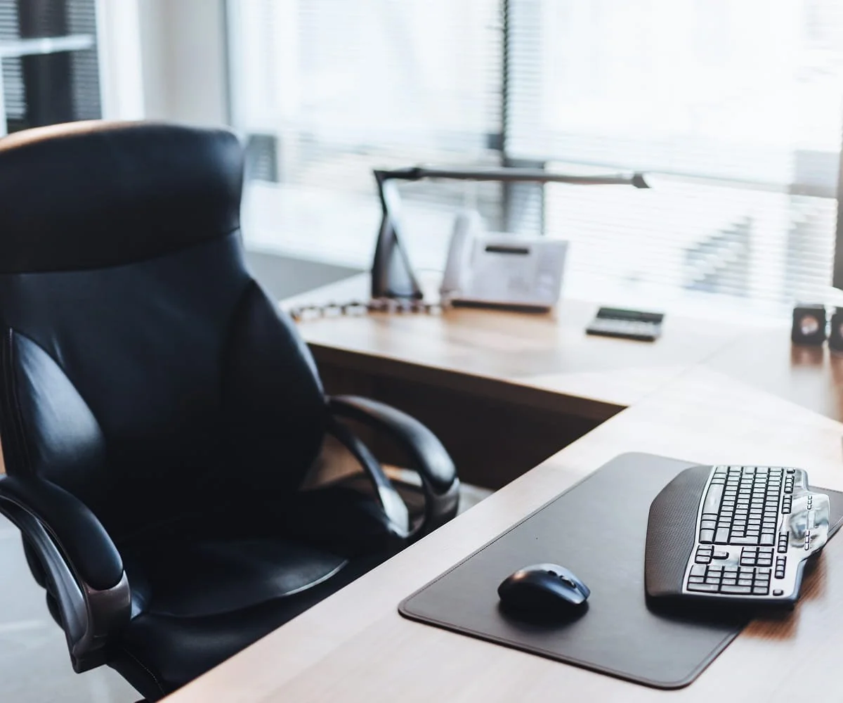 Office desk with a black ergonomic chair, a computer keyboard, mouse, and a mouse pad in a bright room with large windows and blinds.