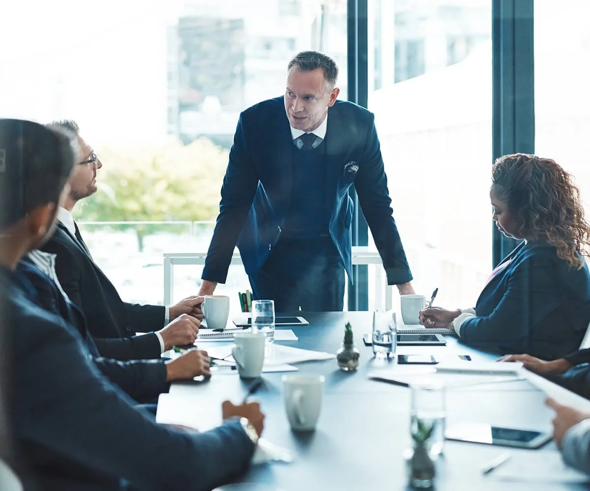 Business meeting in a modern conference room with one man standing and four seated, engaged in discussion, coffee cups and notebooks on the table.