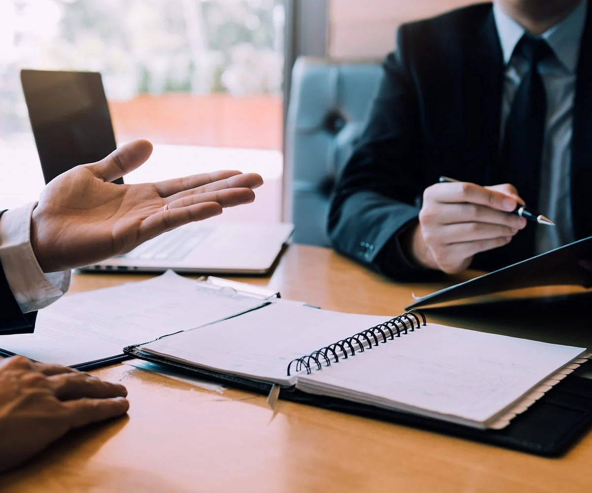 Two people having a business discussion at a wooden table with notebooks, documents, and a laptop, one person gesturing with their hand while the other takes notes.