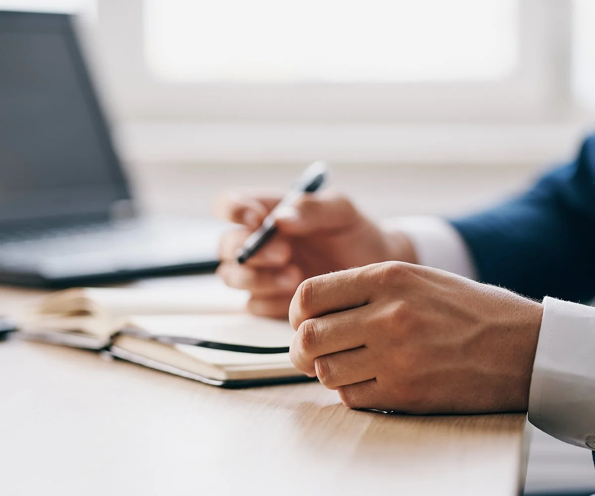 Close-up of a person in a suit holding a pen and writing on a notebook at a desk with a laptop in the background.