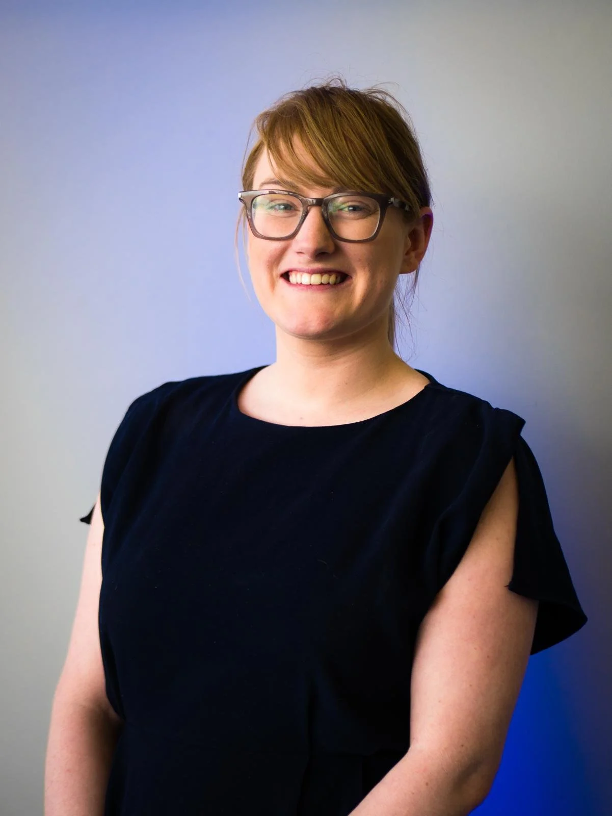A woman with short reddish-brown hair, glasses, and a black top smiling in front of a plain background.