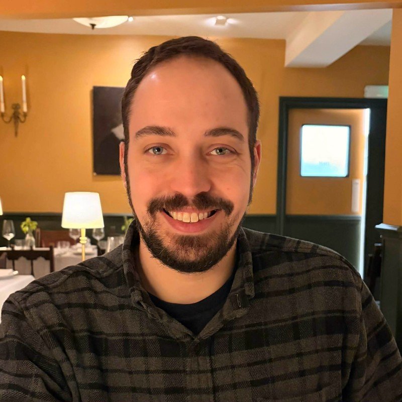 Smiling man with dark hair and a beard in a dimly lit restaurant with yellow walls, white tablecloths, and elegant table settings.