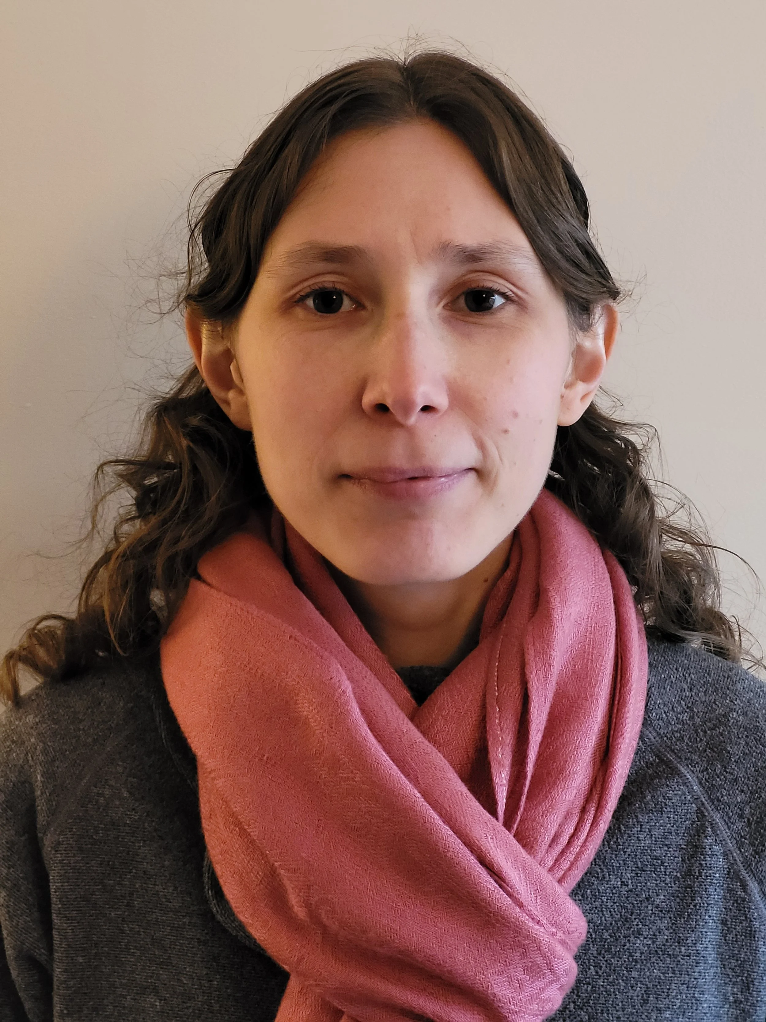 A young woman with shoulder-length wavy dark hair, wearing a pink scarf and a dark gray sweater, looking directly at the camera against a plain beige wall.