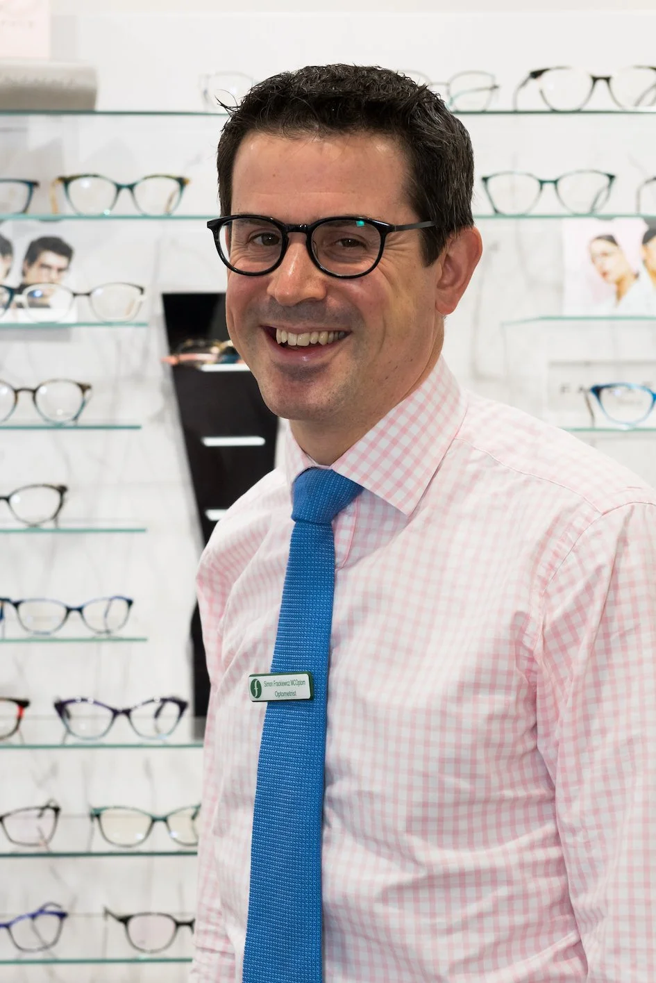 A man with dark hair and glasses smiling, wearing a pink checkered shirt and a blue tie, standing in front of a display of eyeglasses in an optical store.