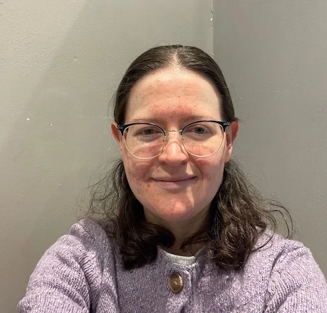 A woman with long brown hair, glasses, and a light purple sweater smiling at the camera against a gray wall.
