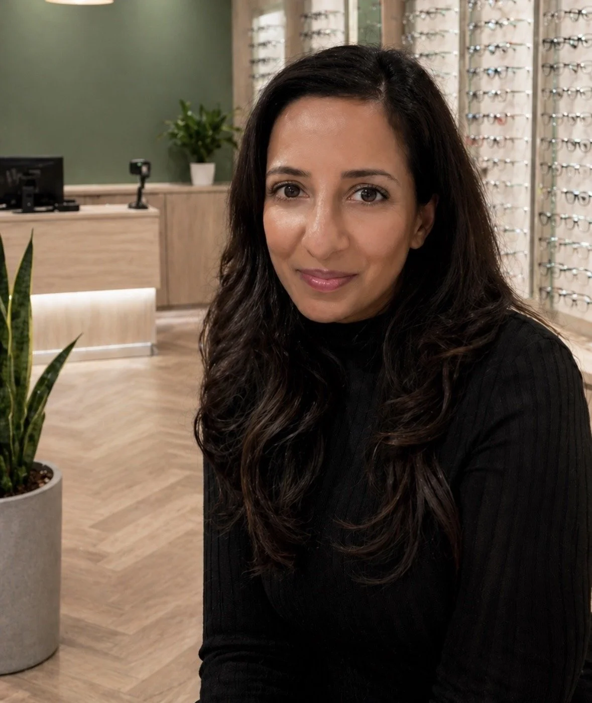 A woman with long, dark, wavy hair and a black turtleneck sweater sitting inside an optical store, with glasses displayed on wall racks in the background.
