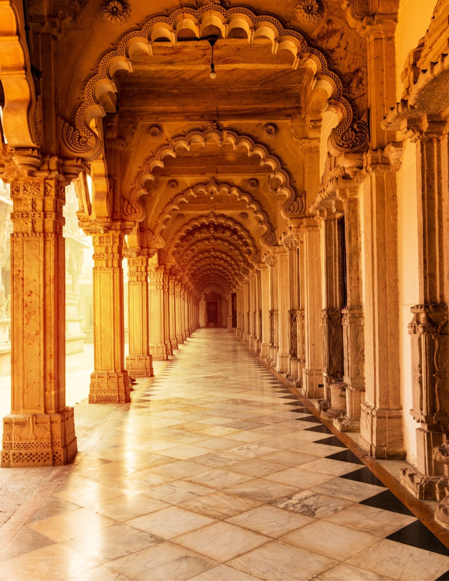 A long corridor with arched ceilings and intricately carved columns made of stone or marble. The floor features a checkered pattern with alternating light and dark tiles, and the lighting casts a warm golden glow.