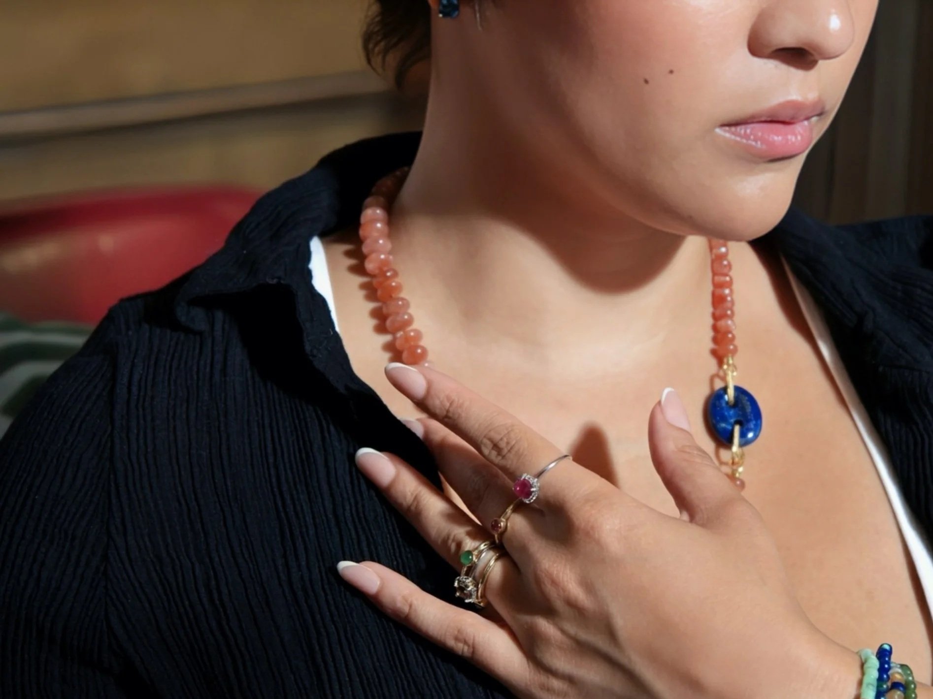Close-up of a person wearing multiple jewelry pieces, including a coral beaded necklace with a blue pendant, and rings on their fingers. The person's face shows part of the cheek, lips, and nose, with short brown hair and a black textured top.