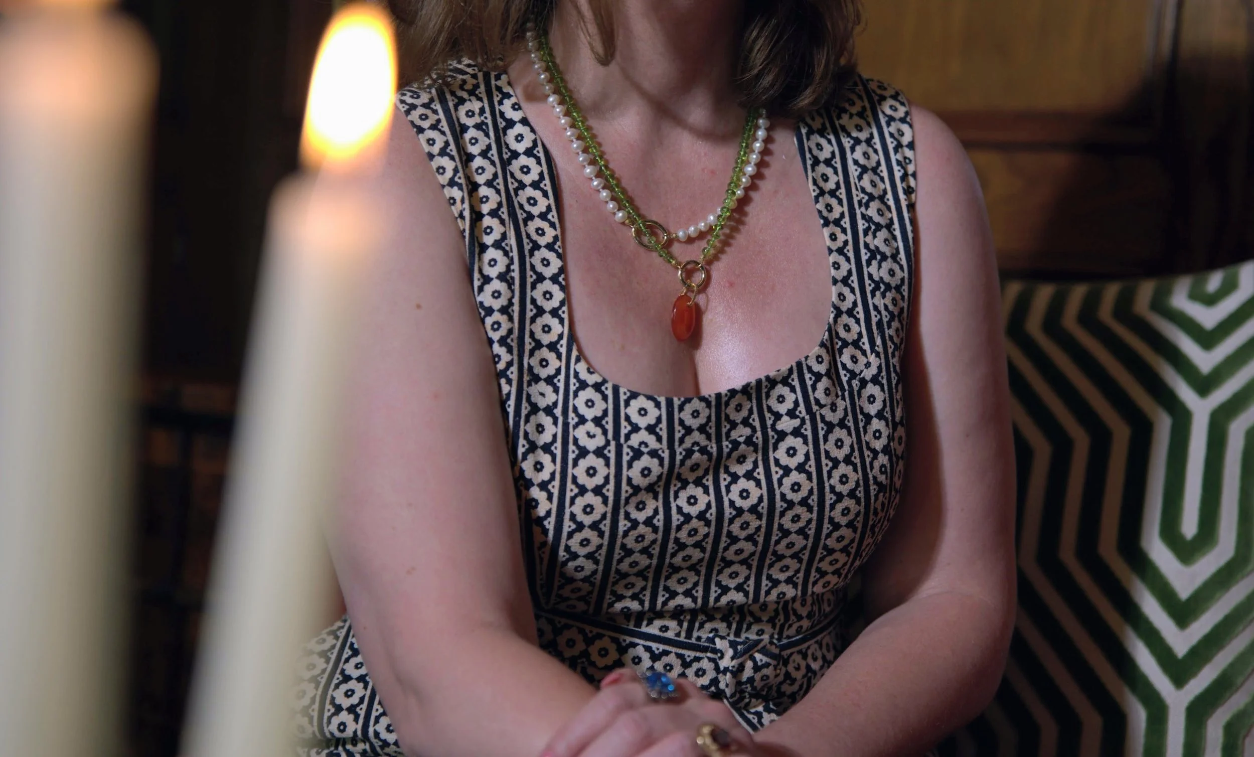 A woman wearing a patterned sleeveless dress and layered necklaces, including one with a large orange pendant, is seated with her arms crossed, in a room with wooden furniture and a green and white zigzag patterned pillow.