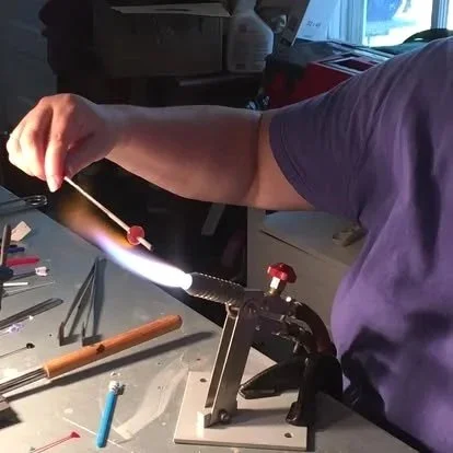 A person in a purple shirt is torch soldering a circuit board at a workbench with tools nearby.