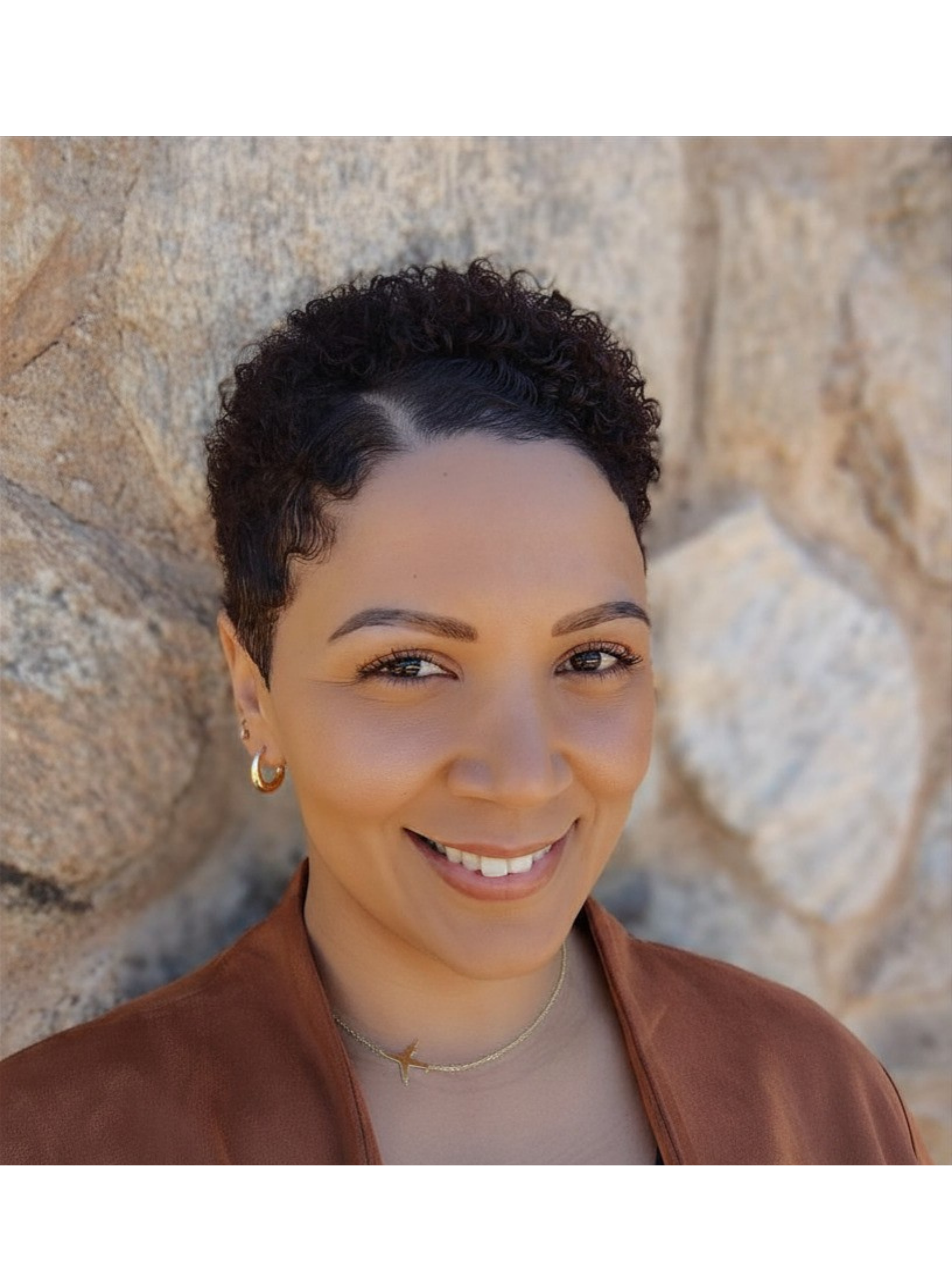 Close-up of smiling woman with short, curly dark hair, wearing gold hoop earrings, a gold airplane charm necklace, and a brown suede jacket, against a background of beige textured stone wall.