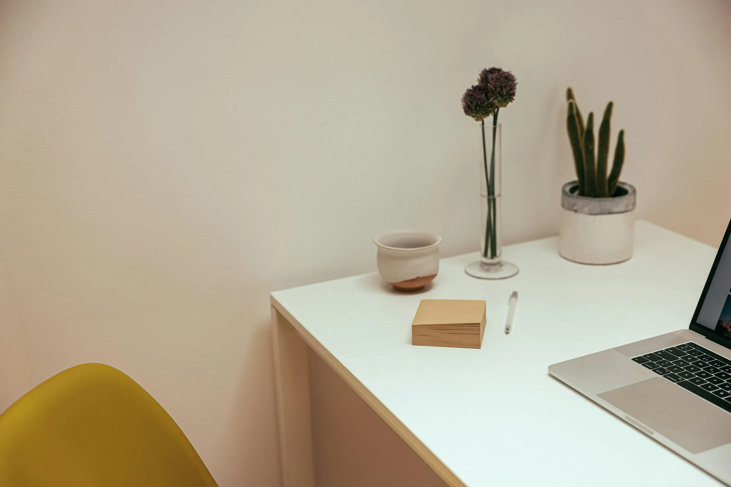 A minimalist white desk with a laptop, a wooden block, a pen, and a potted plant with thick green leaves. Behind the desk, there is a clear vase with purple flowers, a small beige pot, and a yellow chair partially visible in the foreground.