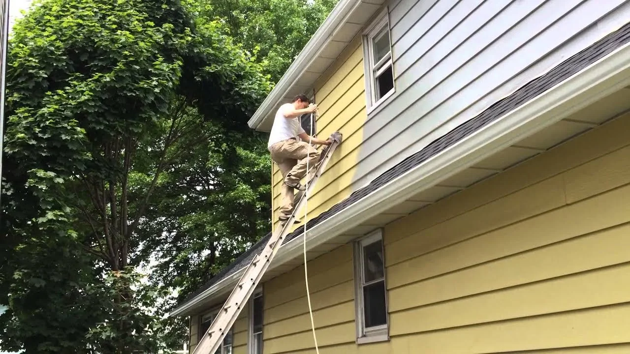 A person working on the exterior of a yellow house, using a ladder and a harness for safety, with trees in the background.