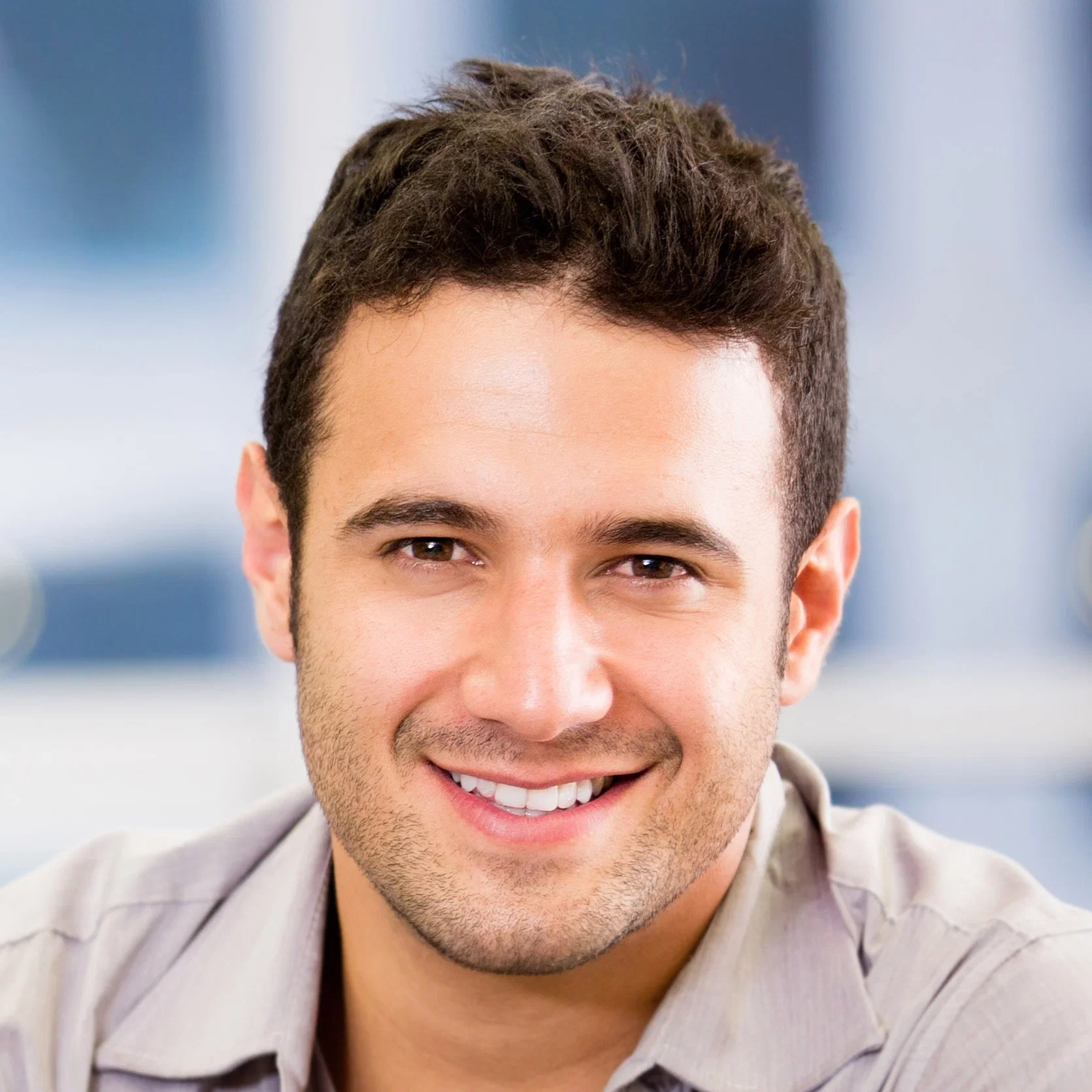 Close-up of a smiling young man with short dark hair, brown eyes, and light facial hair, wearing a light-colored shirt, against a blurred background.