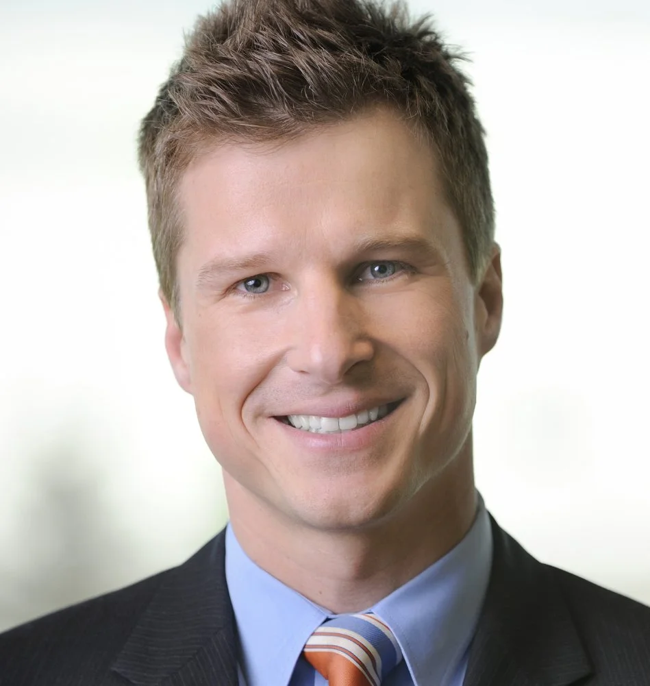 Professional headshot of a smiling young man in a suit and tie with a blurred office background.