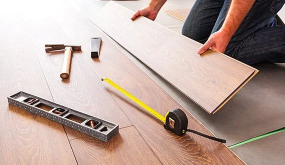 Person installing flooring with tools including a hammer, level, measuring tape, and plank of wood.