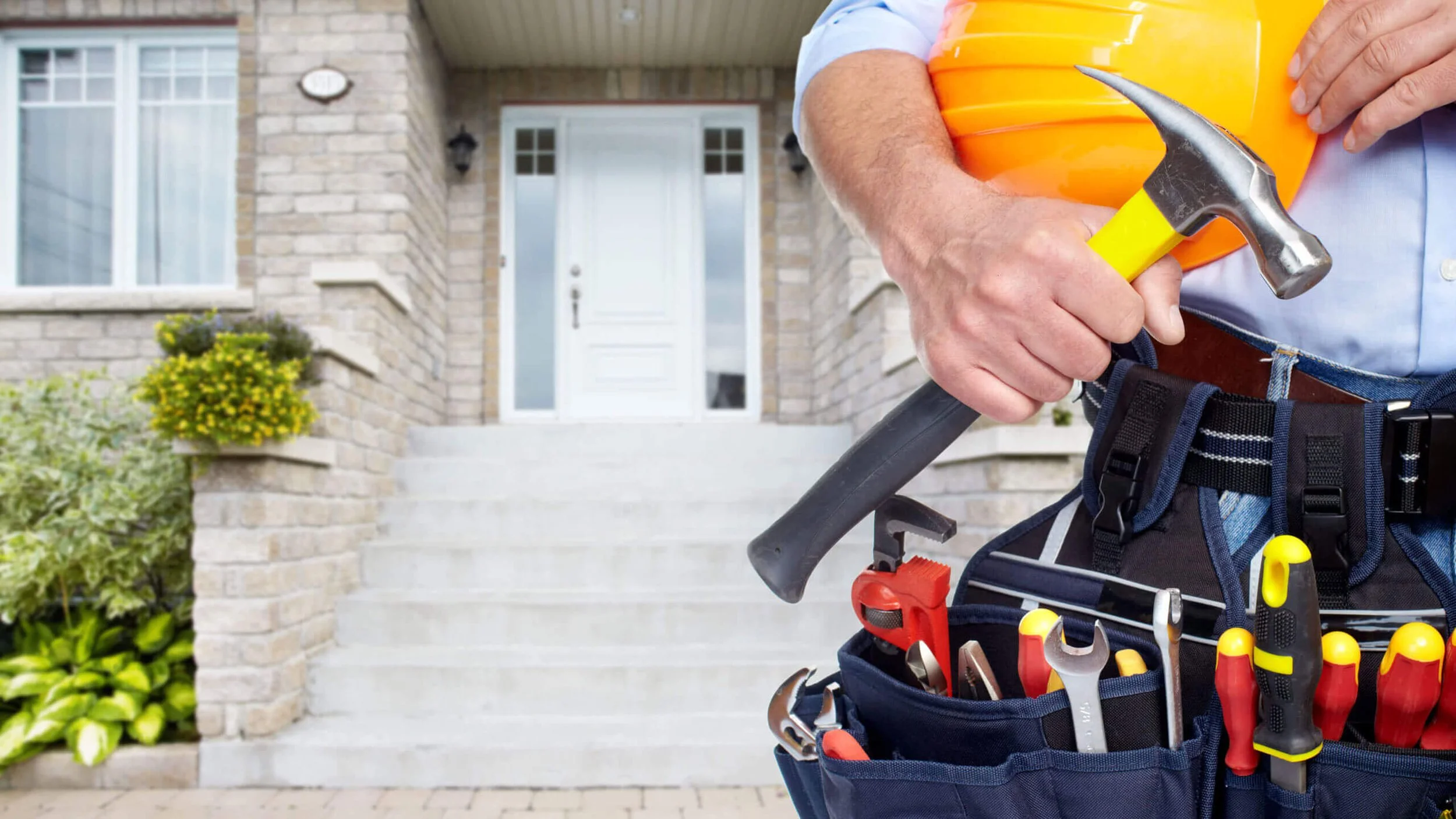 A person holding a hammer and a yellow bucket, wearing a tool belt with various tools in front of a house with steps, a white door, and surrounding greenery.