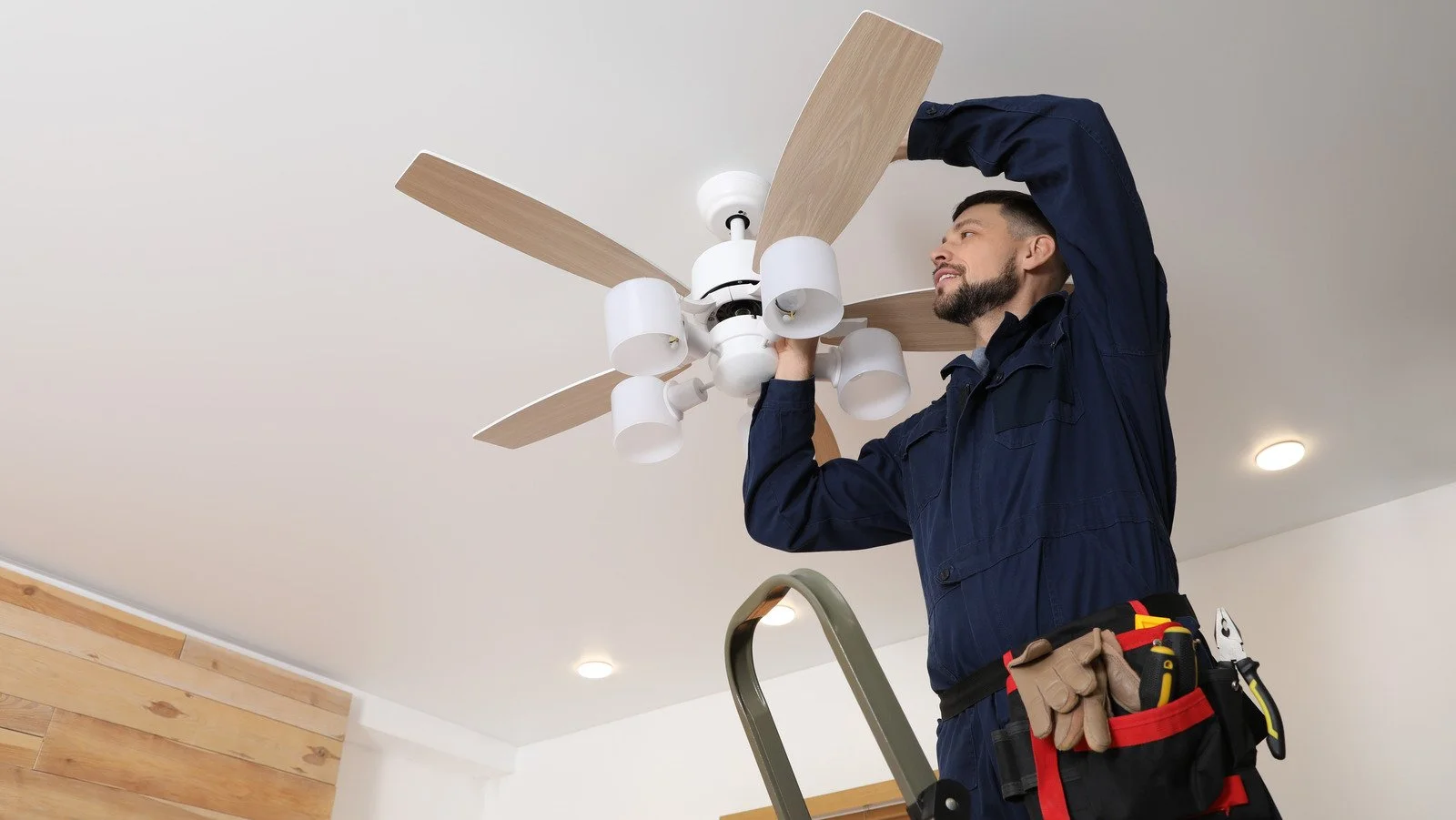 A male electrician fixing a ceiling fan while standing on a step ladder in a room.