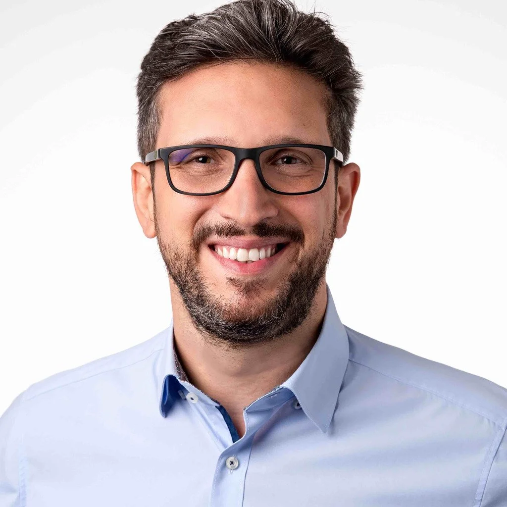 A smiling man with glasses and a beard wearing a light blue dress shirt against a white background.