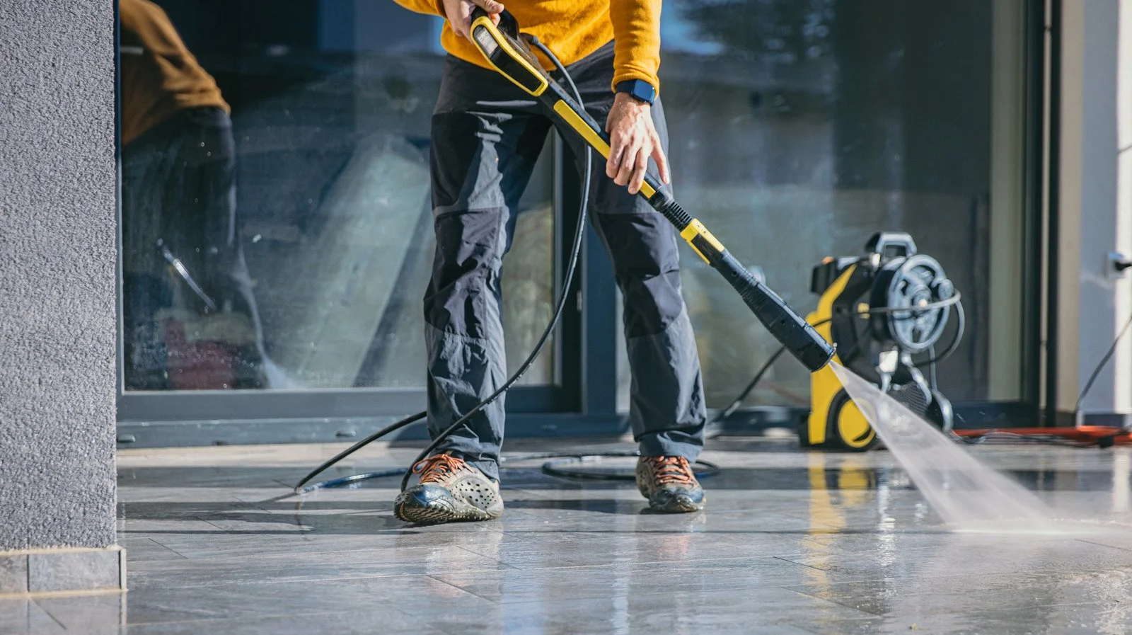 Person pressure washing a tiled floor outdoors near a glass door, with a yellow pressure washer in the background.