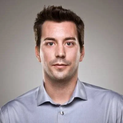 A portrait of a young man with dark hair, wearing a light blue button-up shirt, against a neutral gray background.