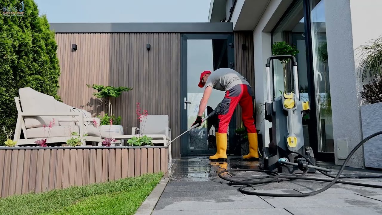 Man pressure washing the patio outside a modern house, wearing yellow boots, red cap, and work clothes.