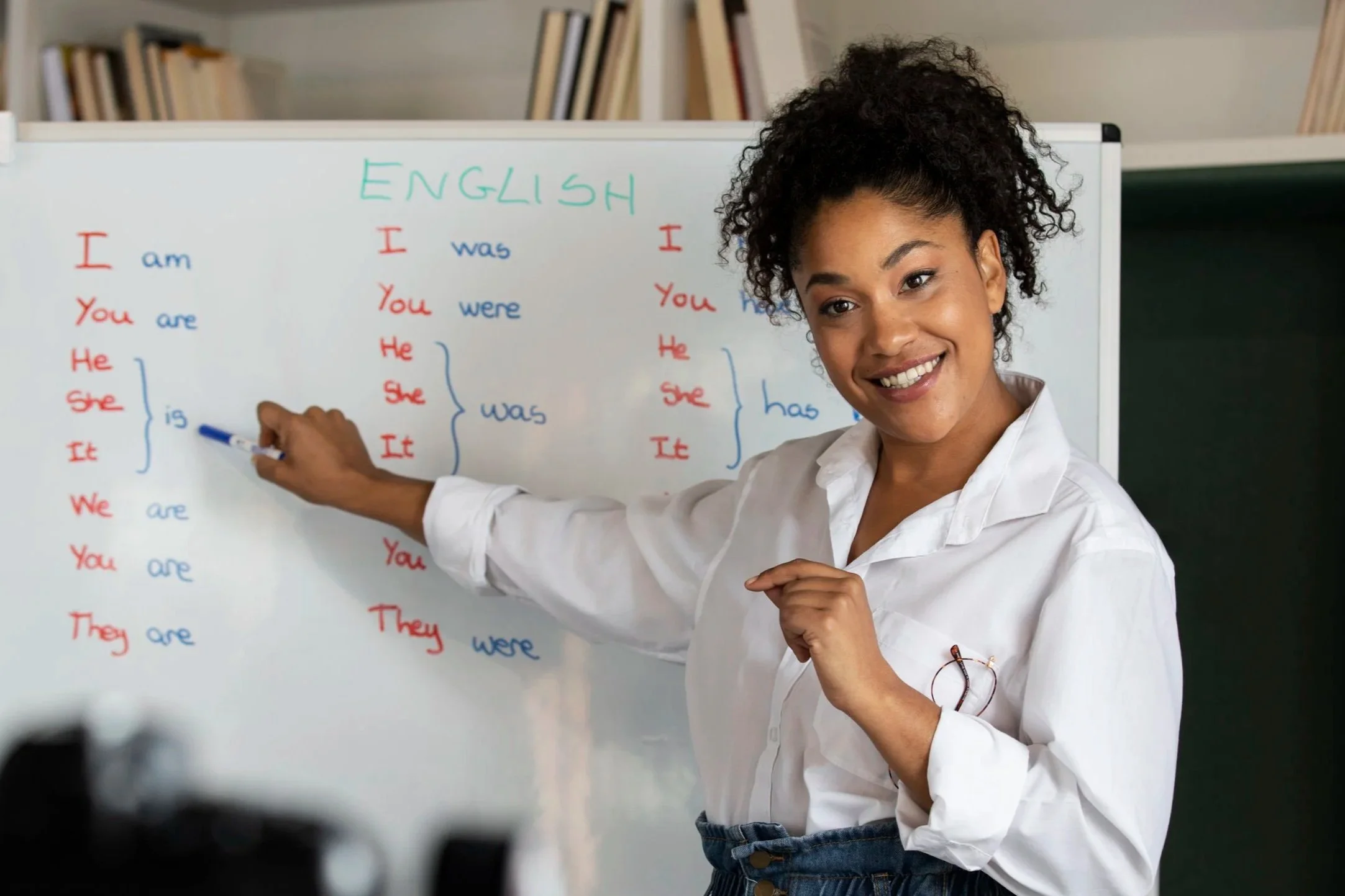 A woman with curly hair smiling and pointing at a whiteboard with English language conjugation notes.